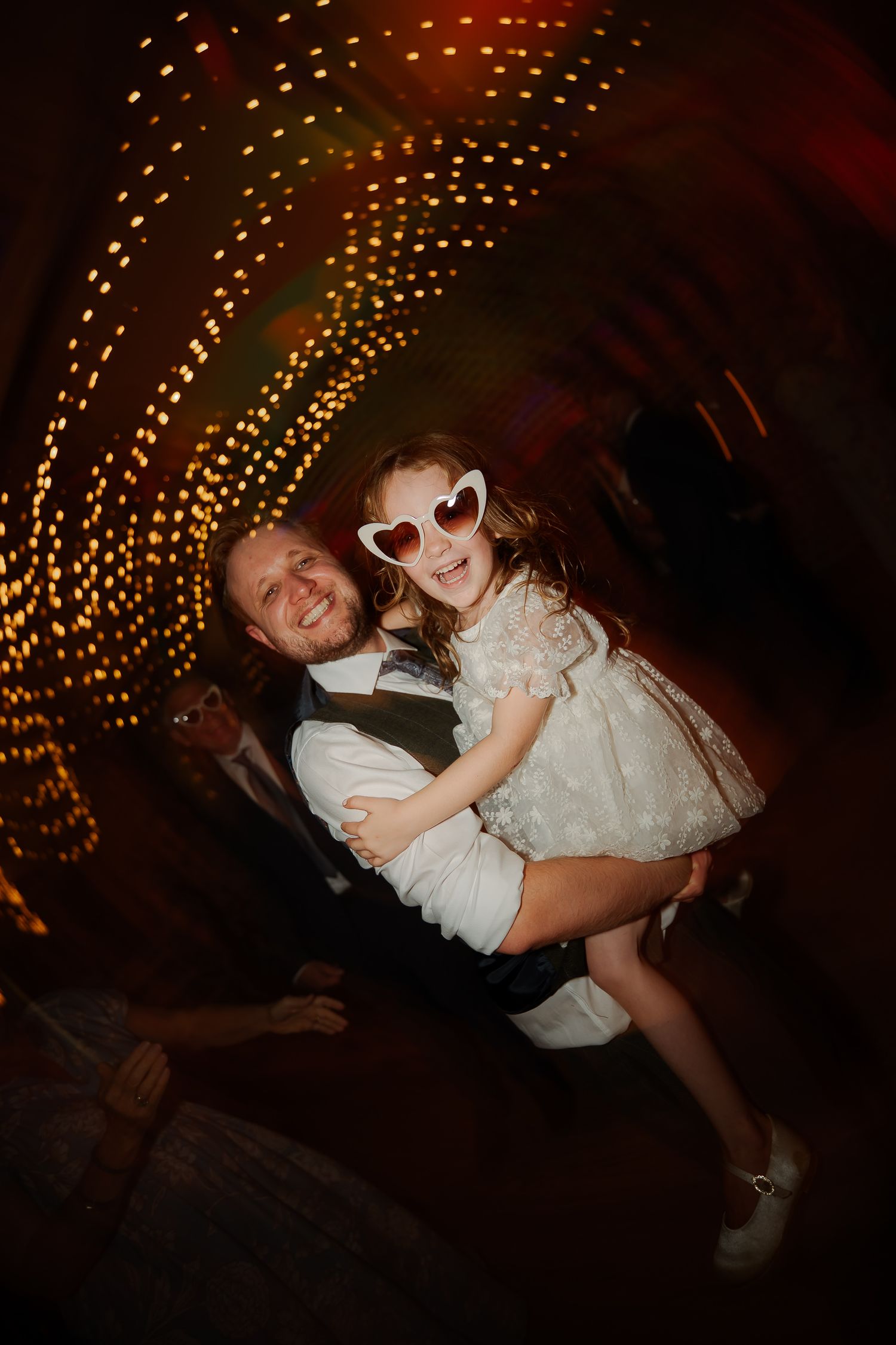 Man holding young girl in heart sunglasses dancing in a glowing light tunnel at a wedding reception.