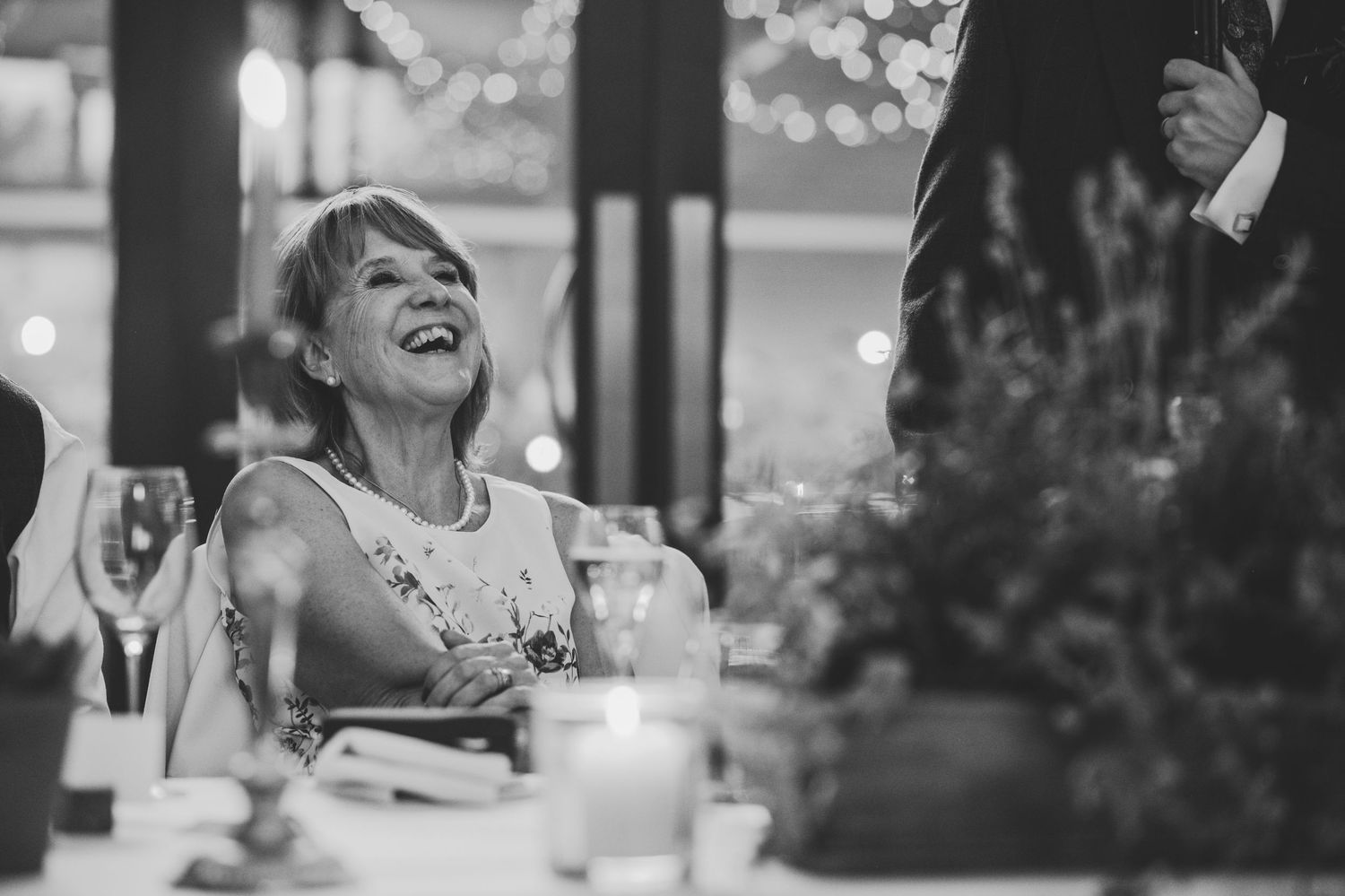 Black and white photo of a laughing woman at a wedding reception dinner table with candles and bokeh lights.