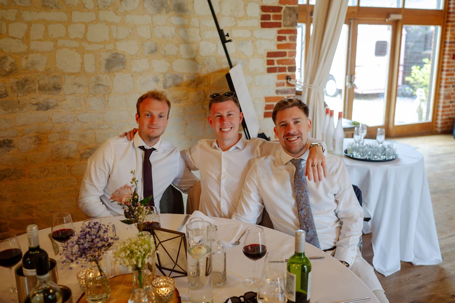 Wedding party in white dress shirts laughs and celebrates together at a rustic stone-walled venue during reception.