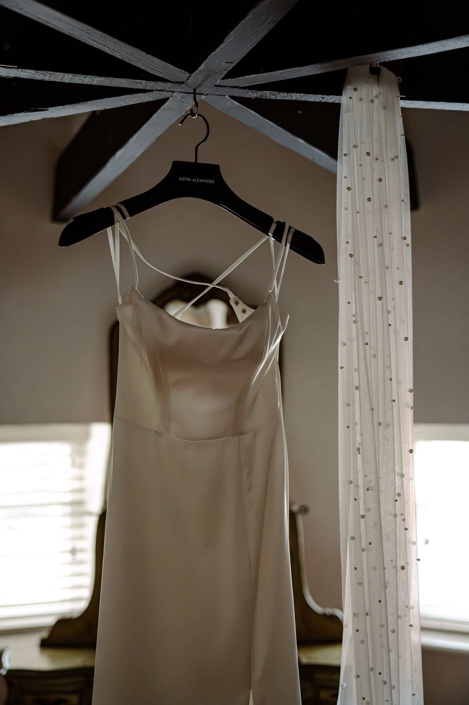 Elegant white wedding dress hangs from wooden beams in natural window light.