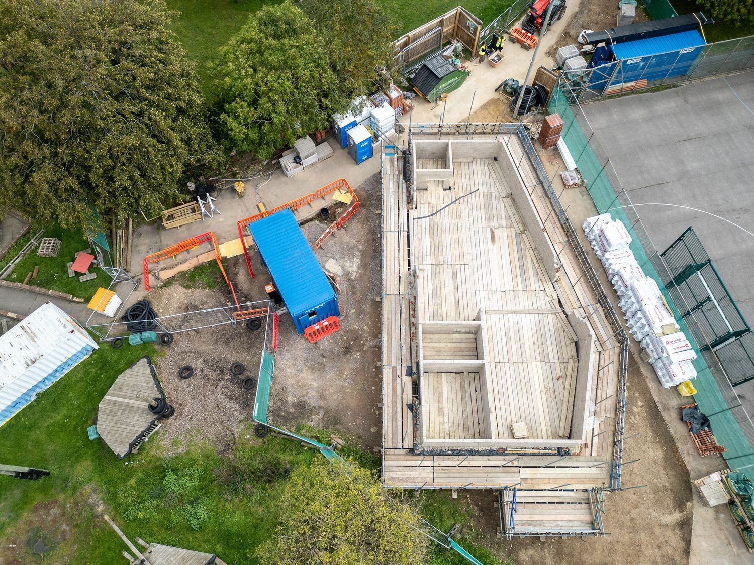 Birds-eye view of construction site showing wooden foundation framing and building materials with blue containers.