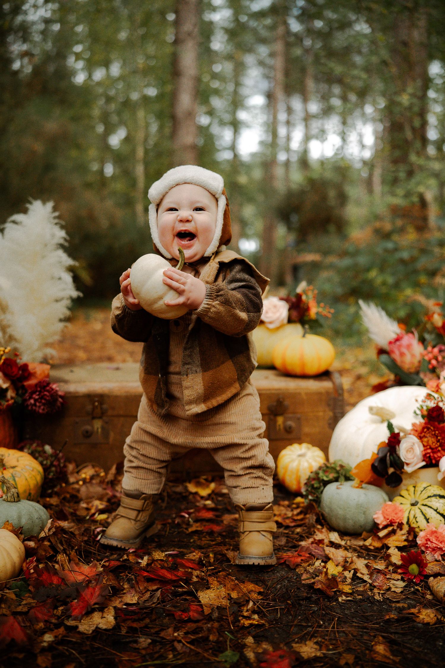 Joyful toddler in autumn outfit holding white pumpkin surrounded by colorful pumpkins and fall foliage in forest setting.
