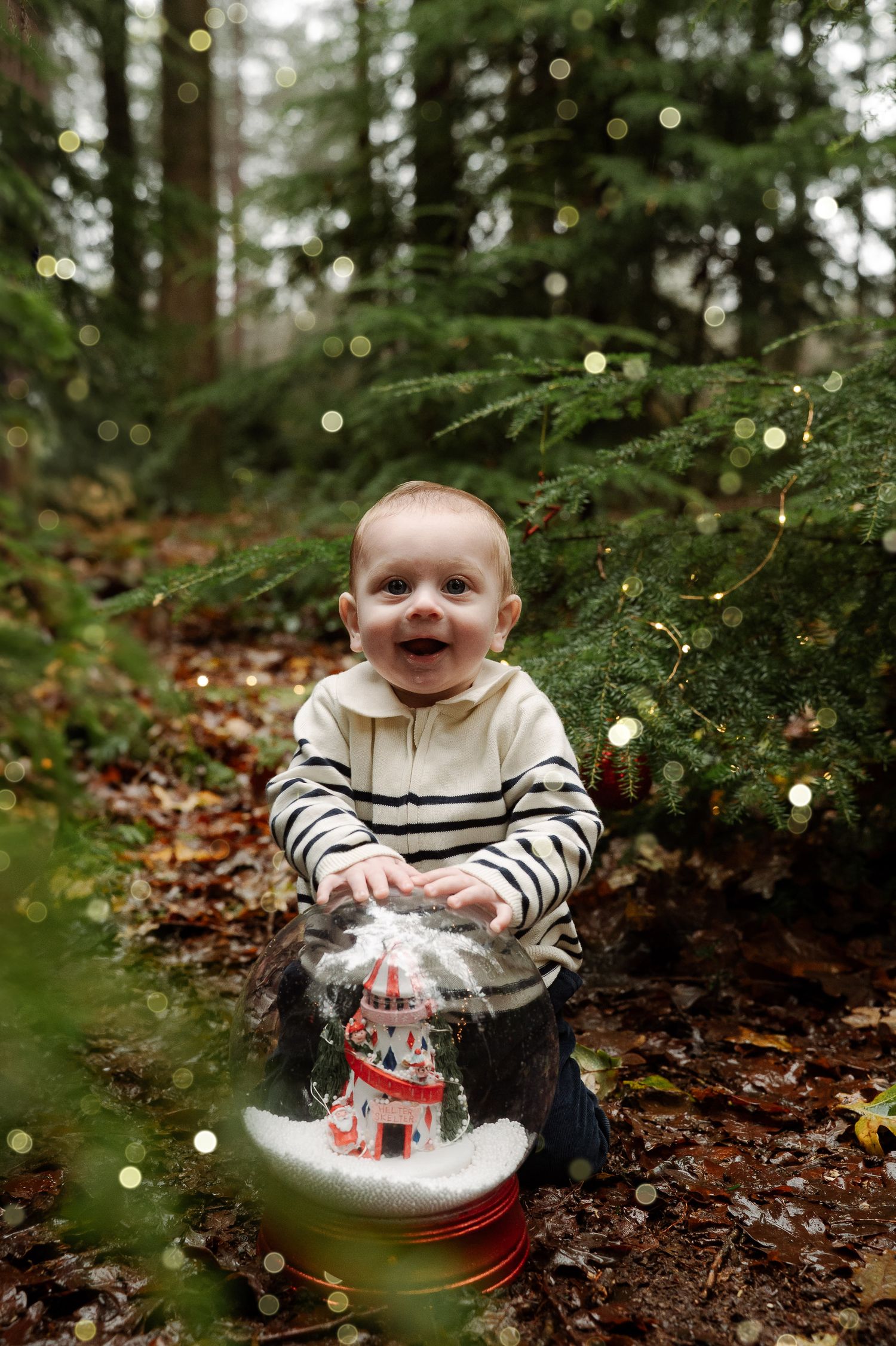 Smiling baby in striped hoodie touching a snow globe ornament in a forest with bokeh Christmas lights.