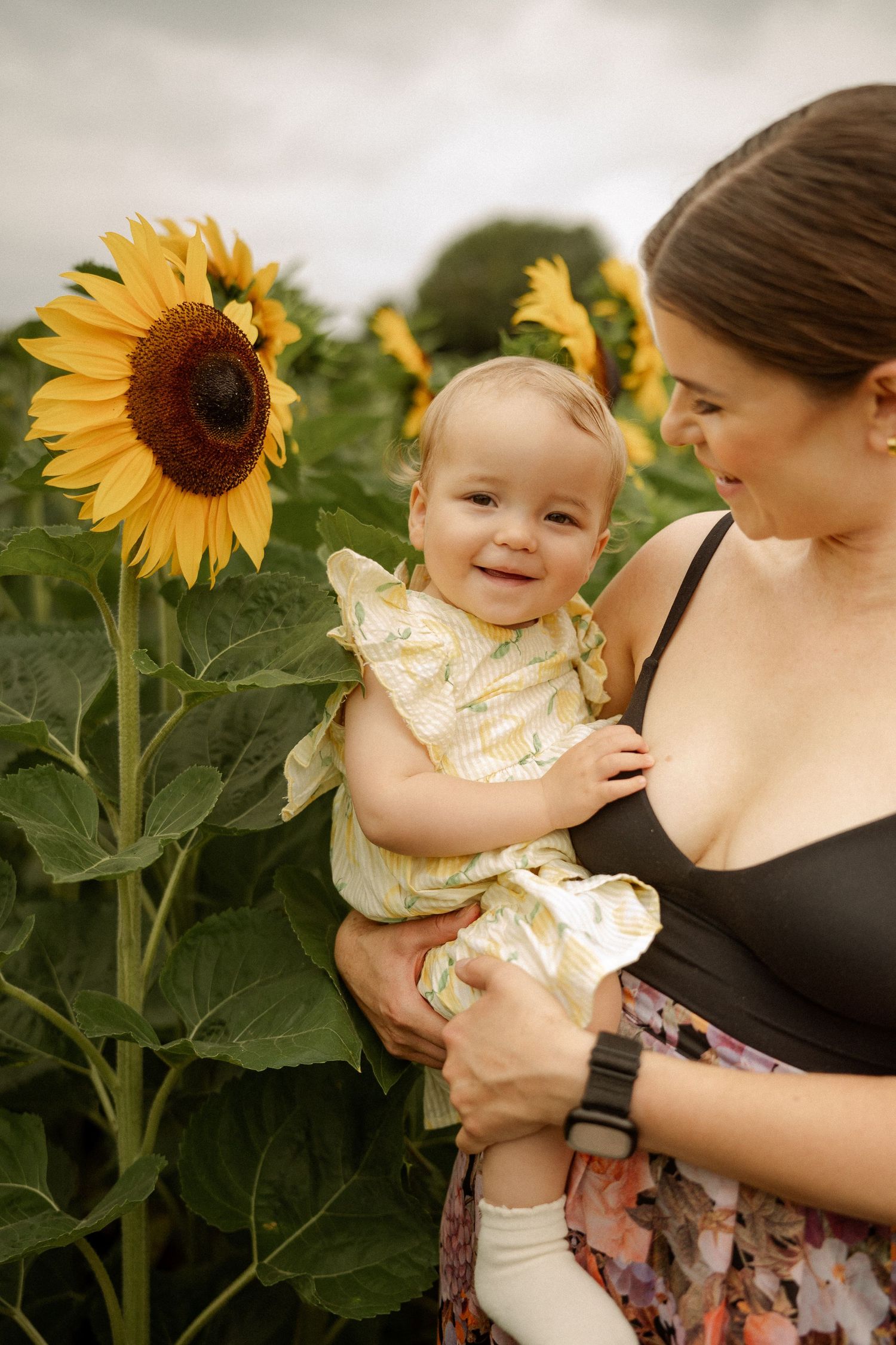 A smiling baby girl in a yellow dress held by her mother in a vibrant sunflower field.