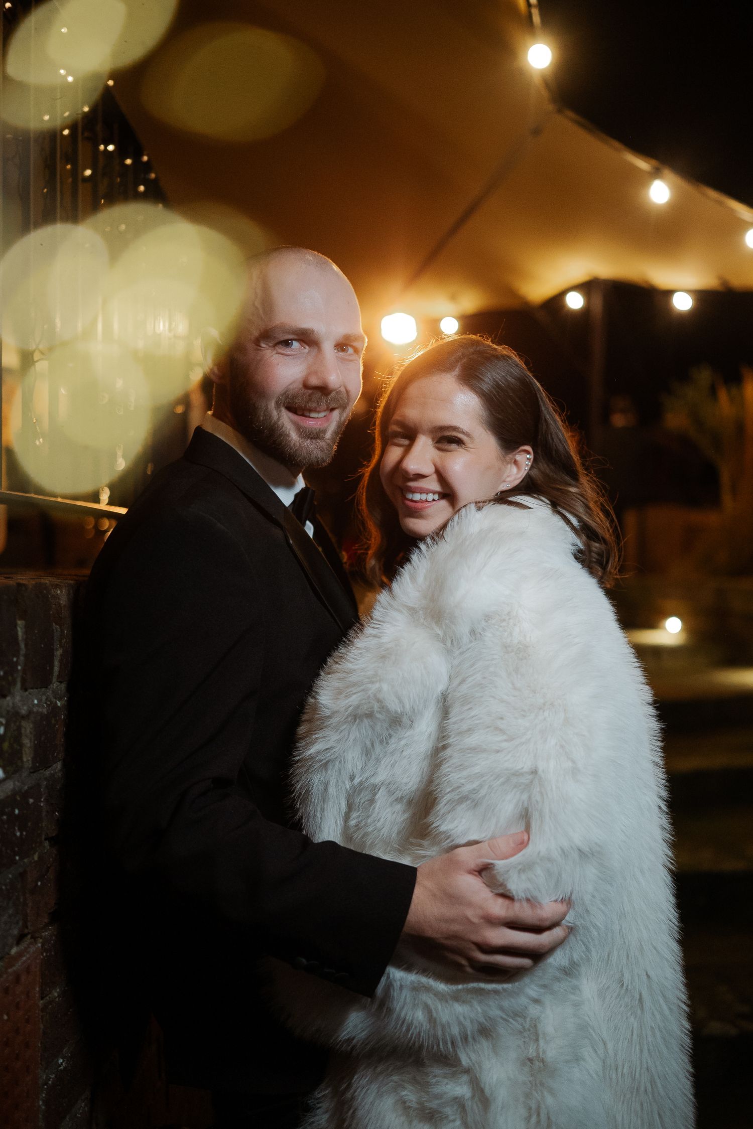 Couple embracing outdoors at night, woman in white fur coat and man in black suit, with warm bokeh string lights behind them.