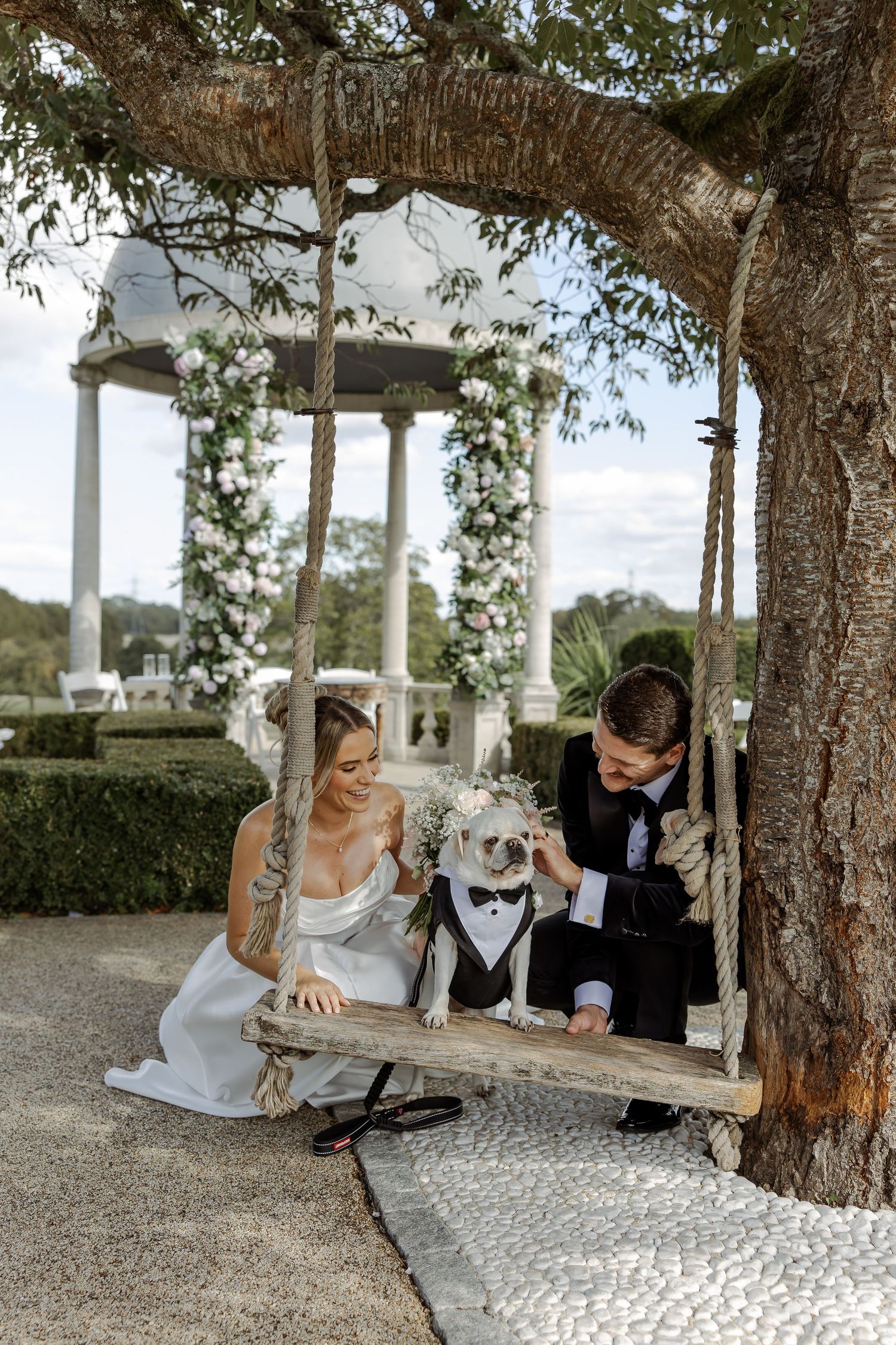 Bride and groom sit on a rustic rope swing with their dog dressed in a tuxedo beneath a floral gazebo.