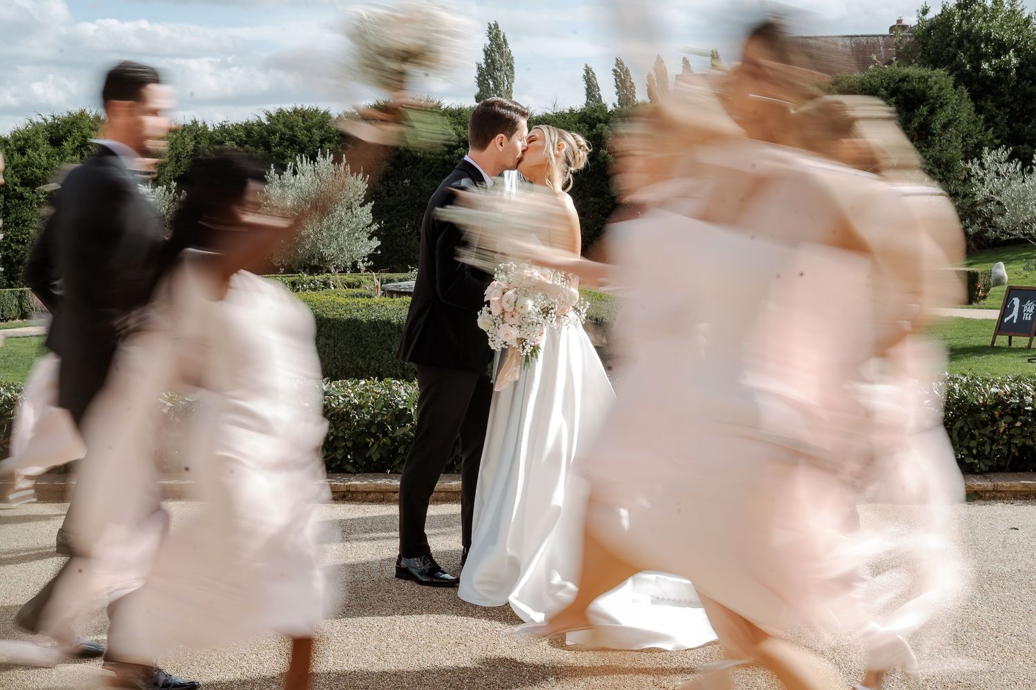 Bride and groom share a kiss surrounded by blurred wedding guests in motion at an elegant outdoor ceremony.