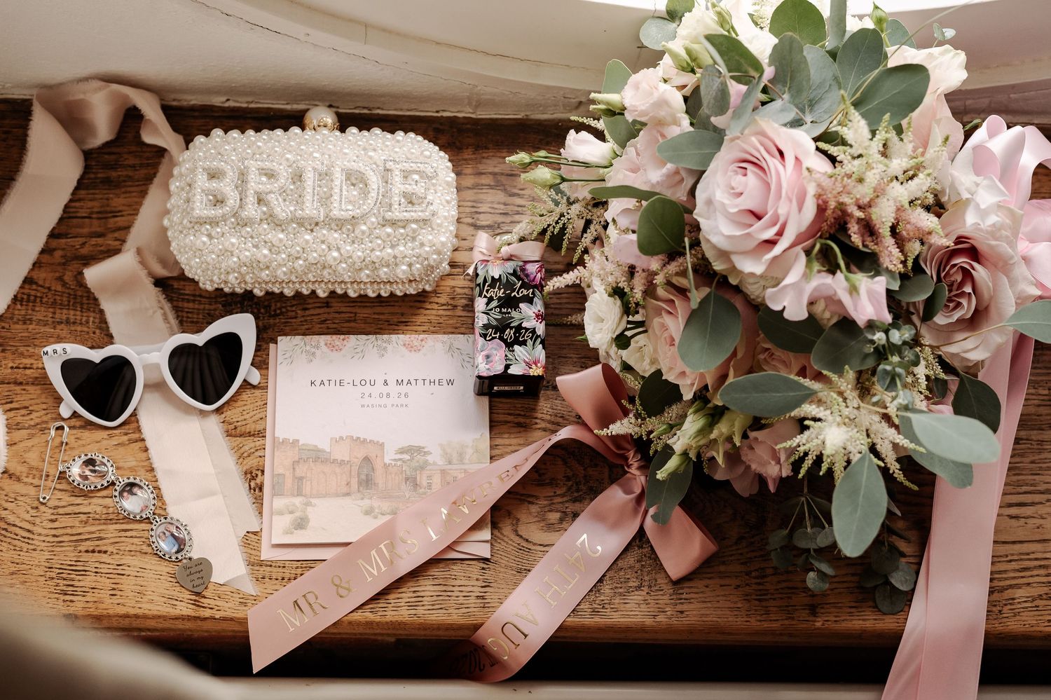 Bridal flat lay featuring a pearl BRIDE clutch, pink rose bouquet, heart sunglasses, and wedding stationery on a wooden surface.