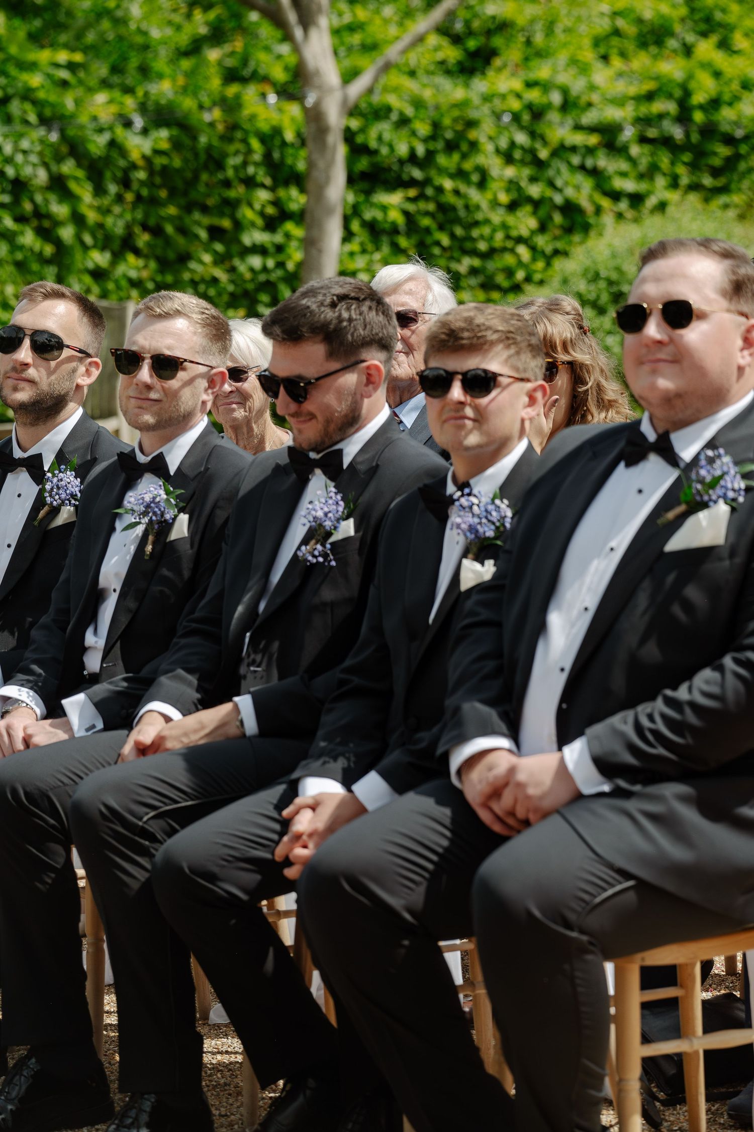 Groomsmen in black suits and sunglasses sit in wooden chairs during an outdoor wedding ceremony.