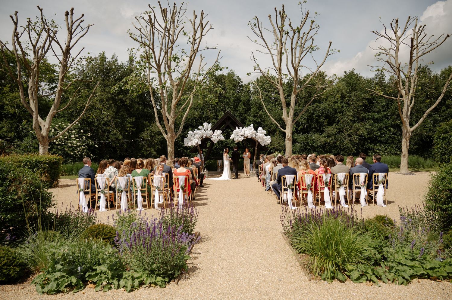 Outdoor wedding ceremony setup with white chairs arranged in rows along a tree-lined path.
