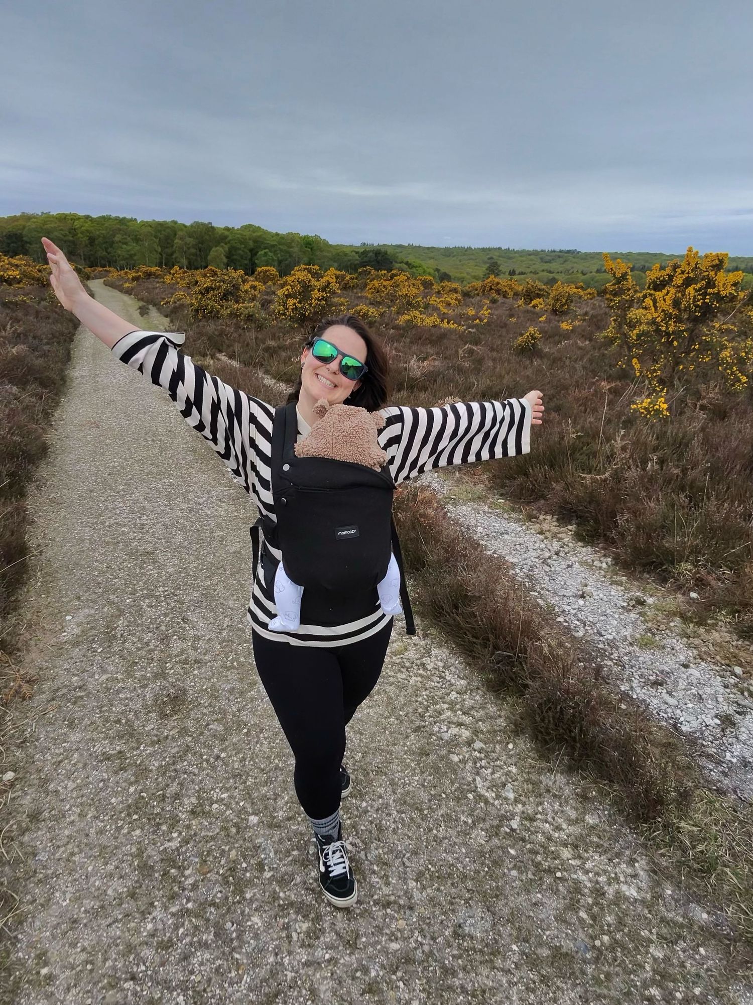 A person in black pants and zebra-striped sleeves walks along a gravel path with gorse bushes on a cloudy day.