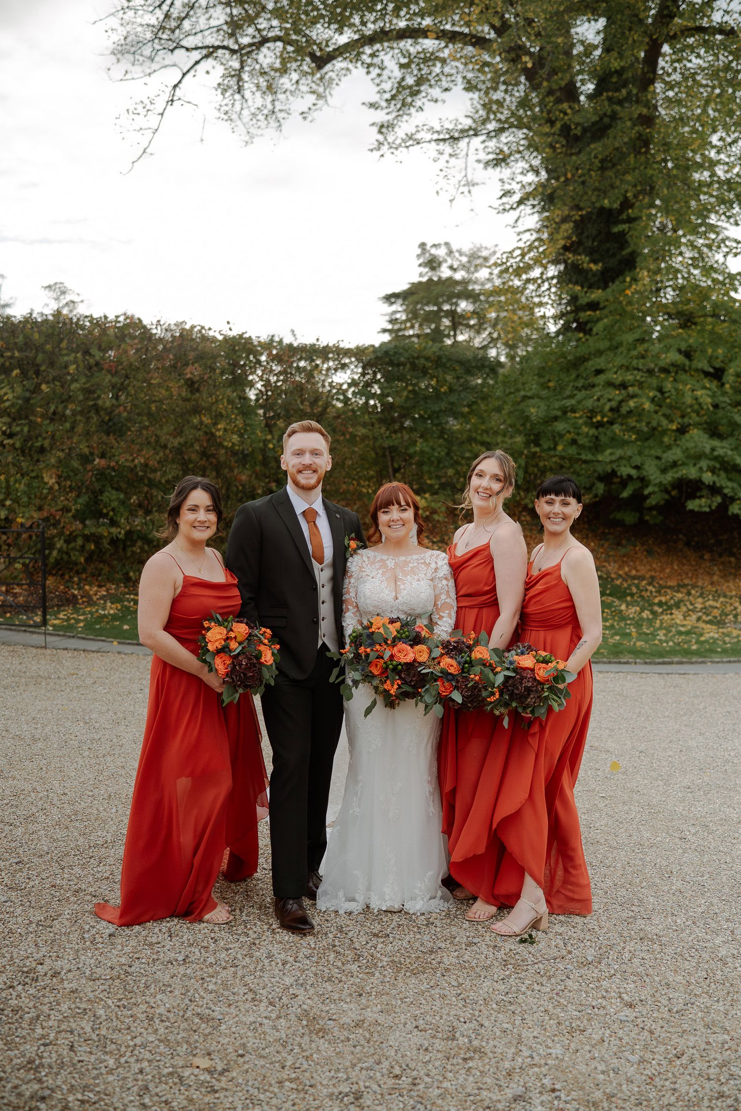 Wedding party in red bridesmaid dresses and formal attire pose together on gravel path during autumn season.