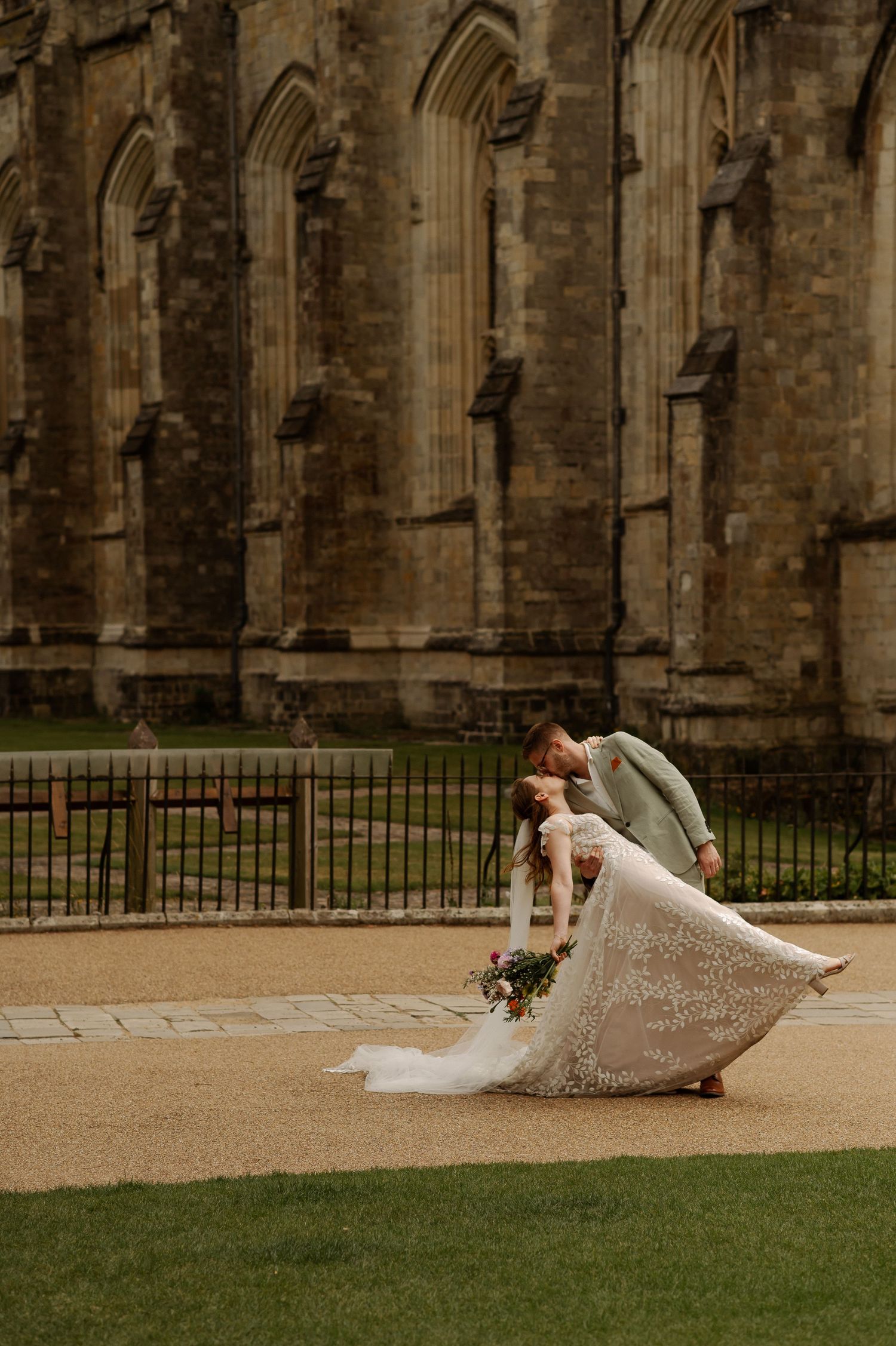 A bride in a lace wedding dress sits gracefully on stone steps in front of a historic Gothic cathedral exterior.