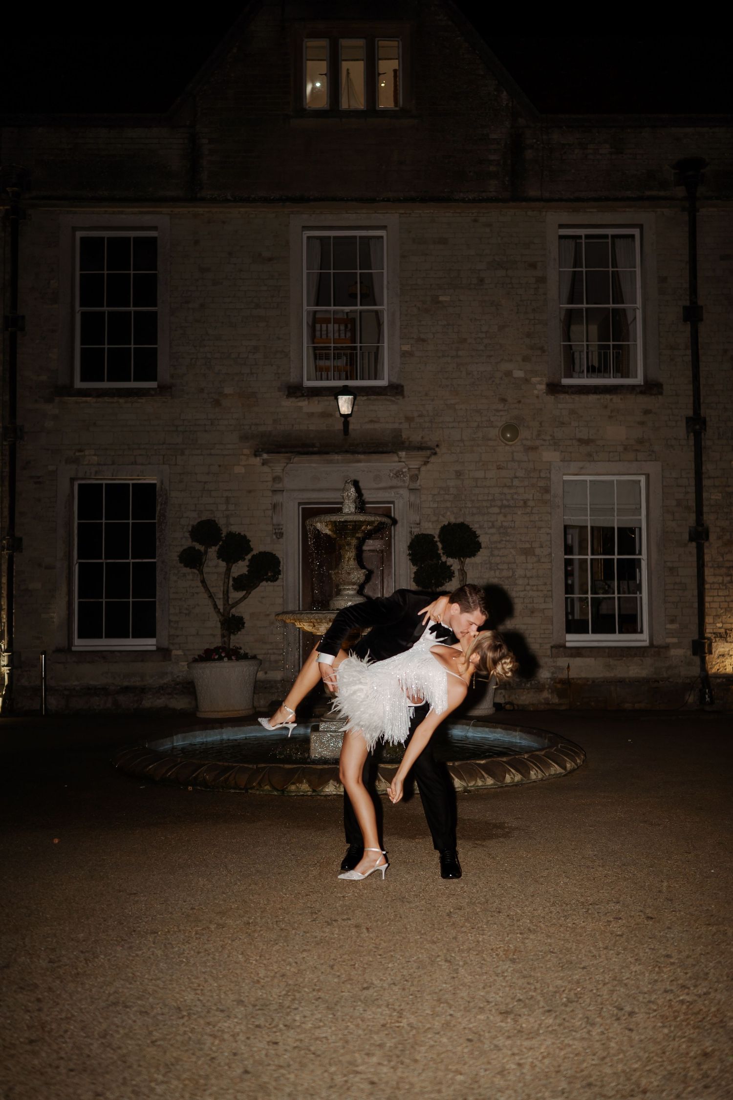 Couple sharing a romantic dip kiss in front of a grand illuminated manor house at night.