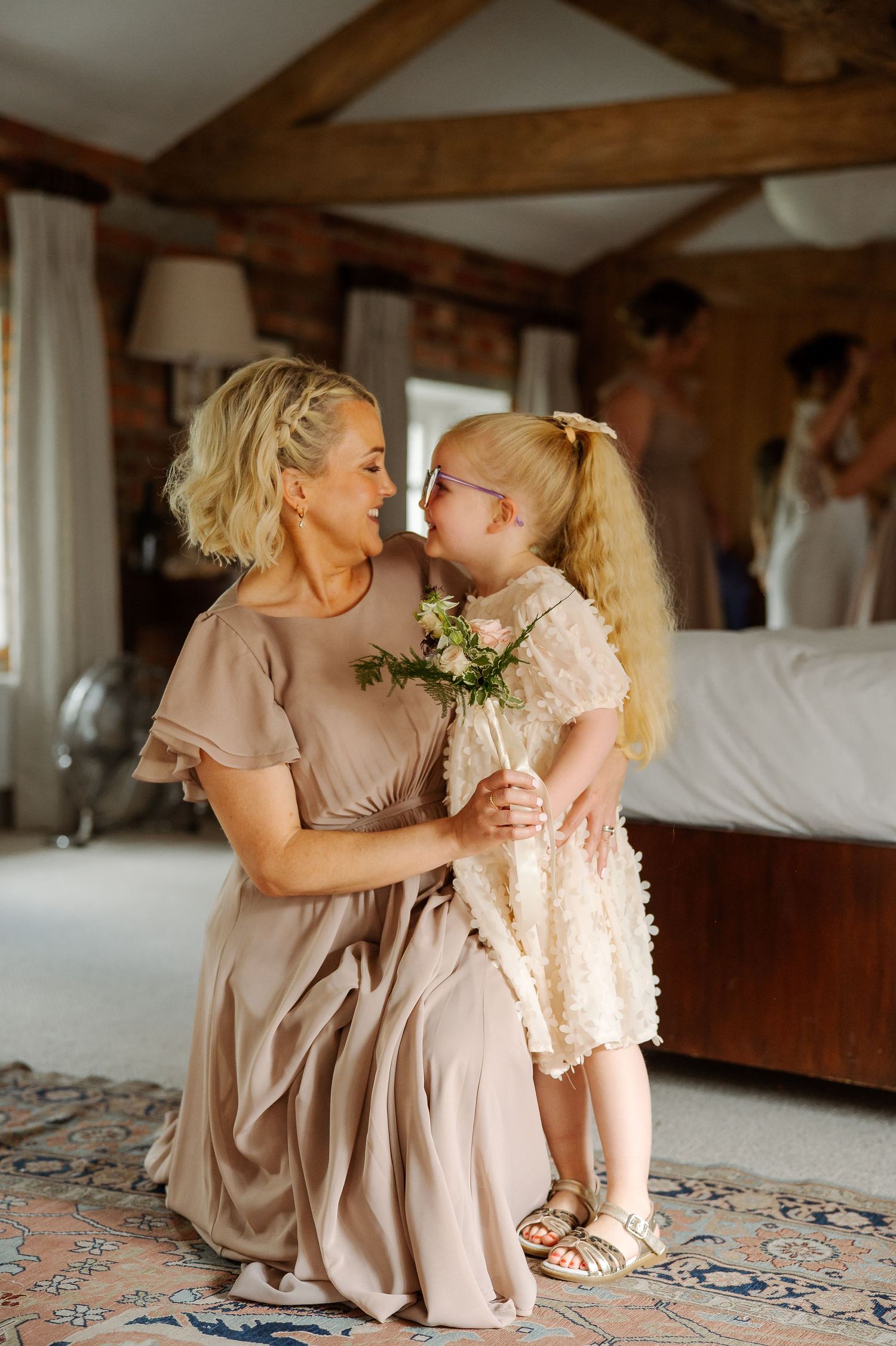 Blonde bridesmaid in taupe dress kneels to embrace young flower girl holding bouquet in rustic wedding venue.