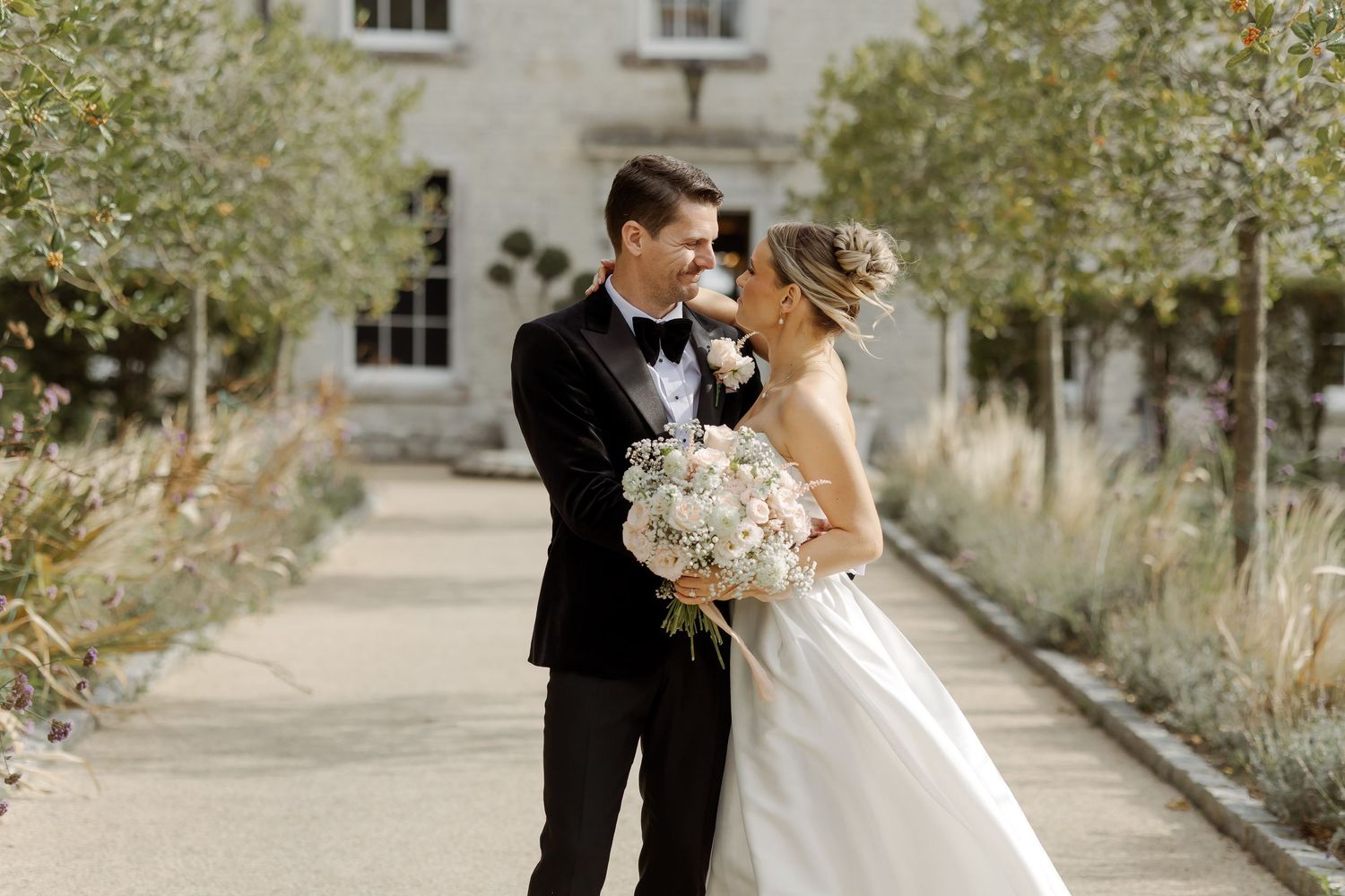 Bride and groom share a romantic moment on a garden path, bride holding a white bouquet, with a elegant manor house backdrop.