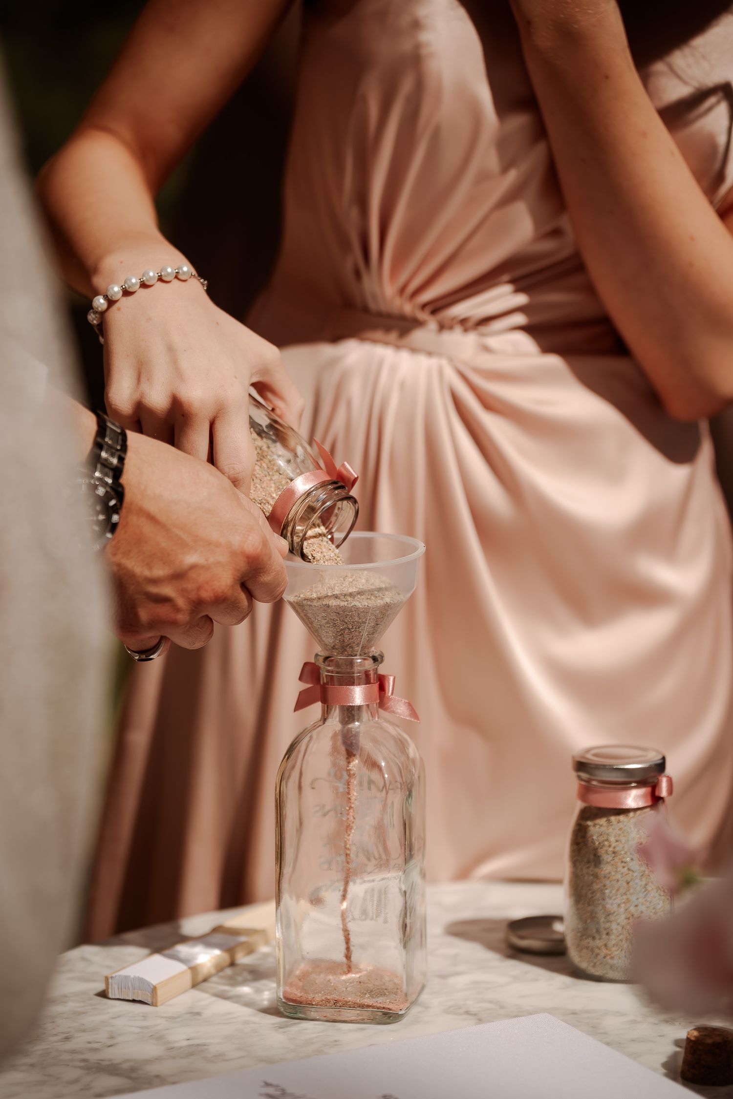 Hands pouring sand into a glass bottle through a funnel during a wedding sand ceremony, with a bridesmaid in pink.