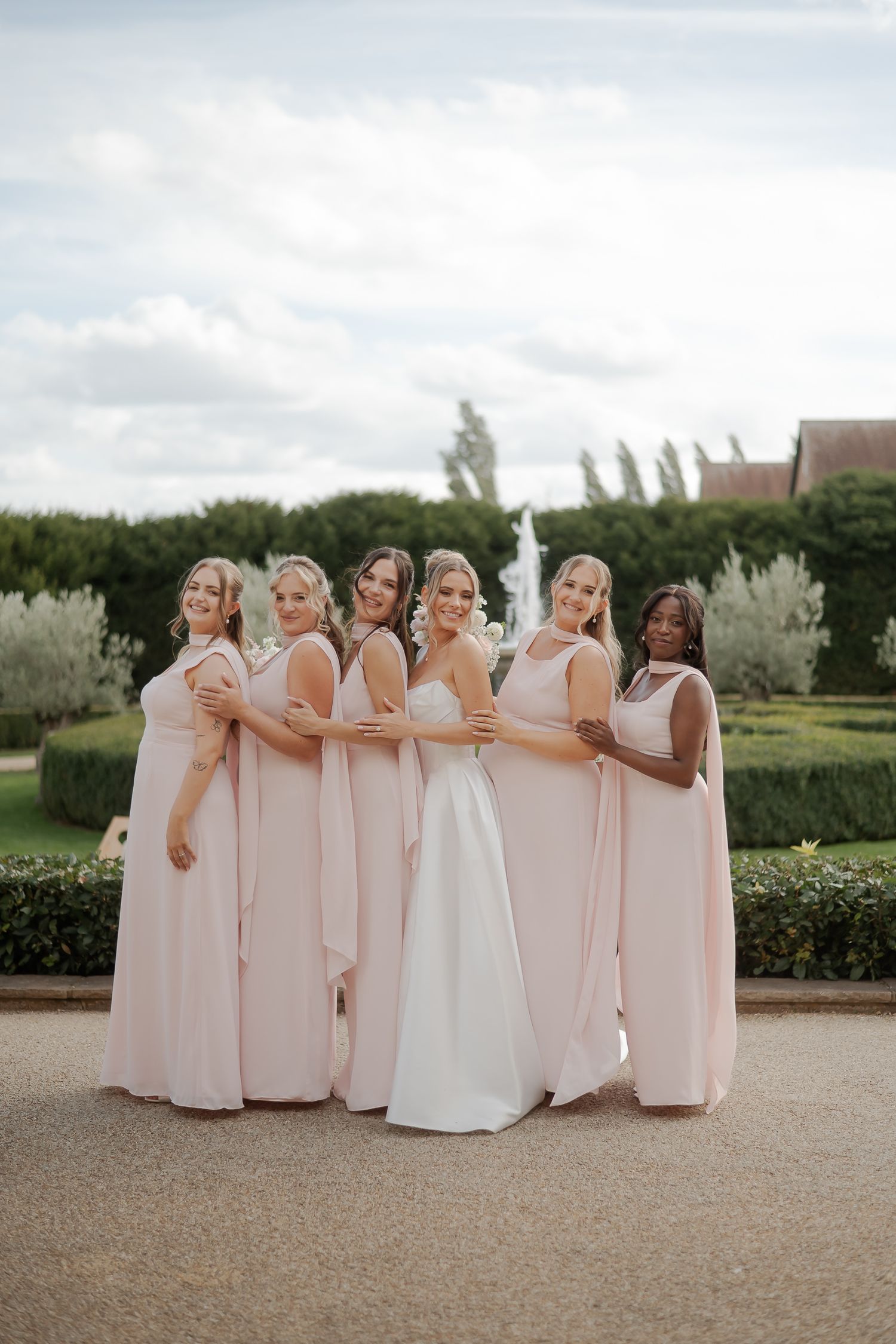 Bride in white gown poses with five bridesmaids in blush pink dresses in a lush garden setting.