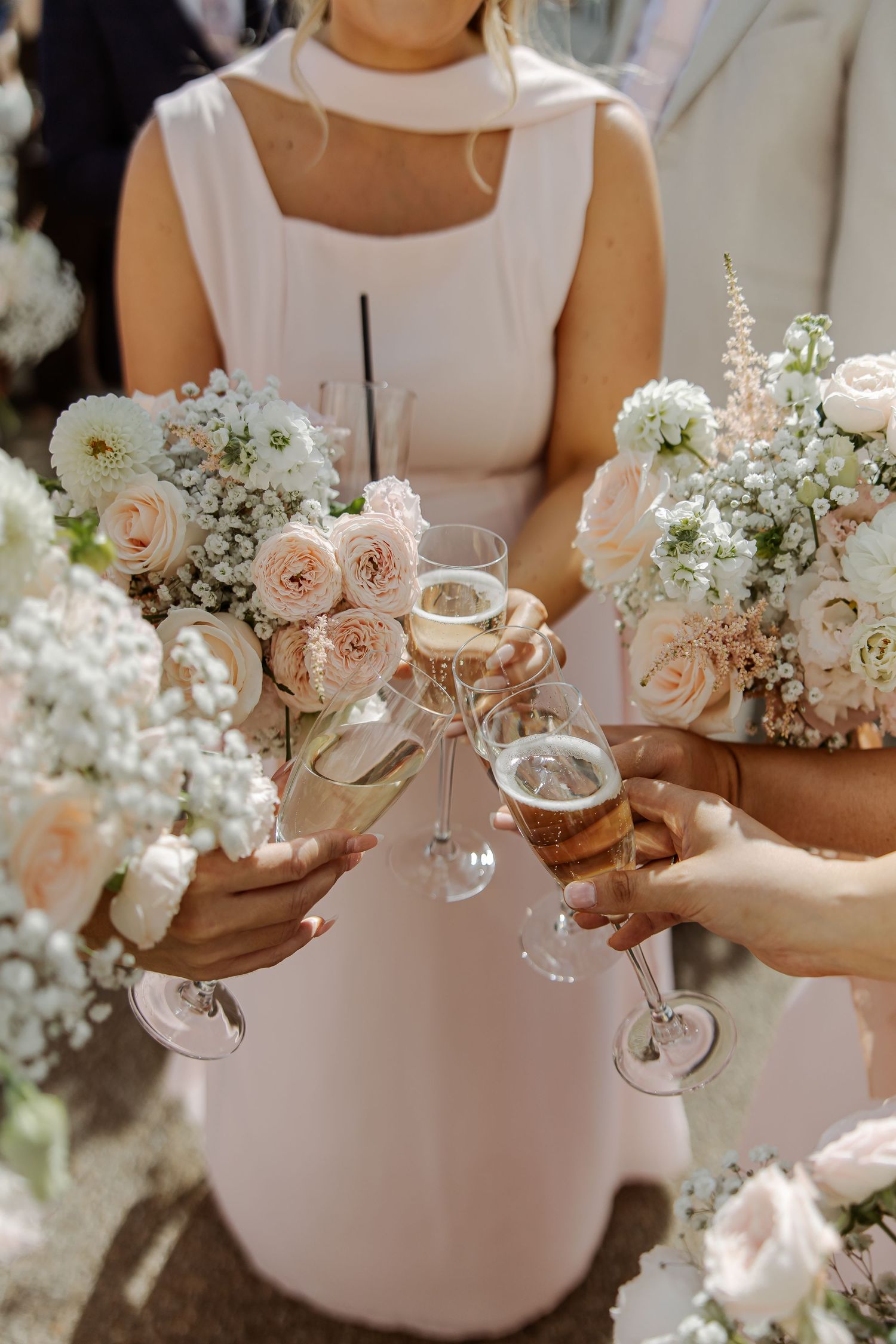 Bridesmaids in blush dresses toasting with champagne glasses while holding elegant white and pink rose wedding bouquets.