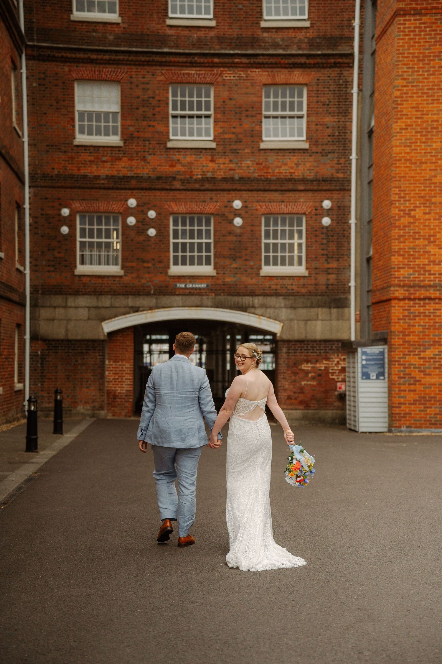 A couple in wedding attire walks away from a brick building with white windows on a cloudy day.