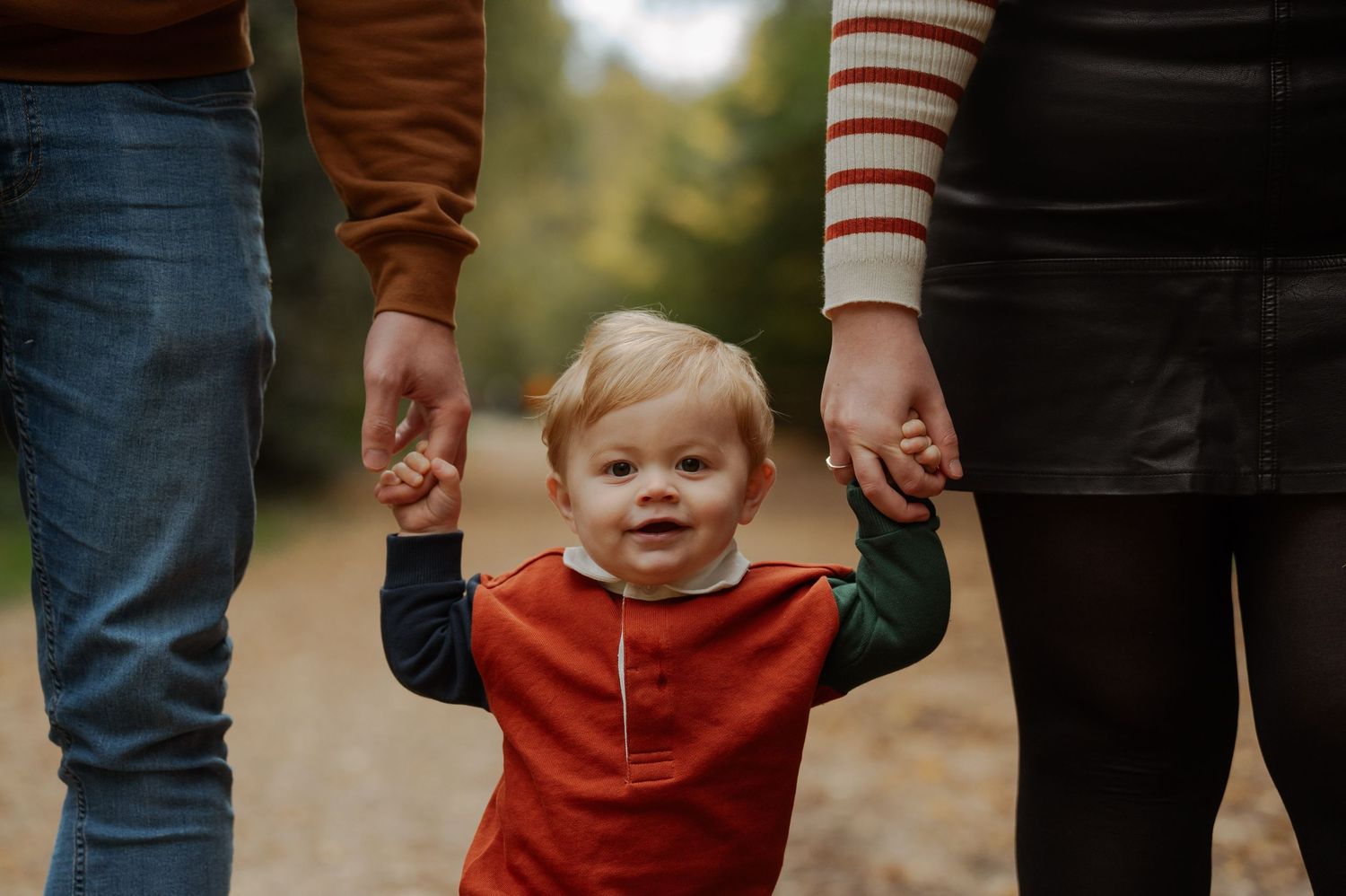 A toddler in a red shirt smiles while holding hands with two adults on a path during autumn.