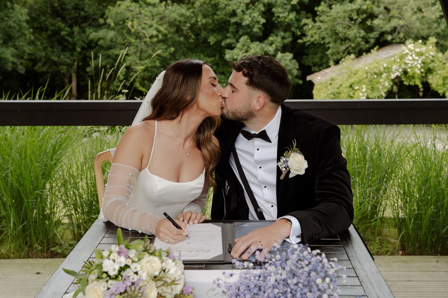 A couple shares a romantic kiss at an outdoor table decorated with purple and white floral arrangements.