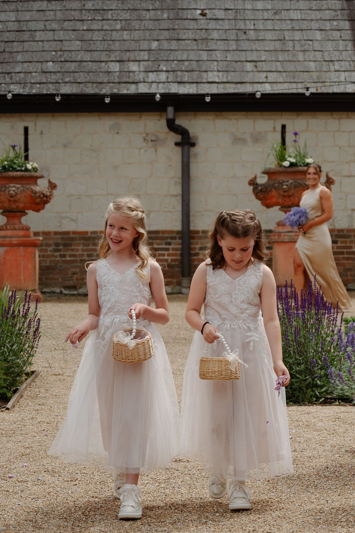 Young flower girls in white dresses carrying baskets walk along a gravel path.