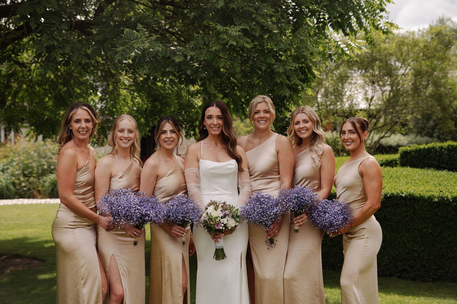 Wedding party wearing champagne-colored dresses poses outdoors holding purple floral bouquets in a garden setting.