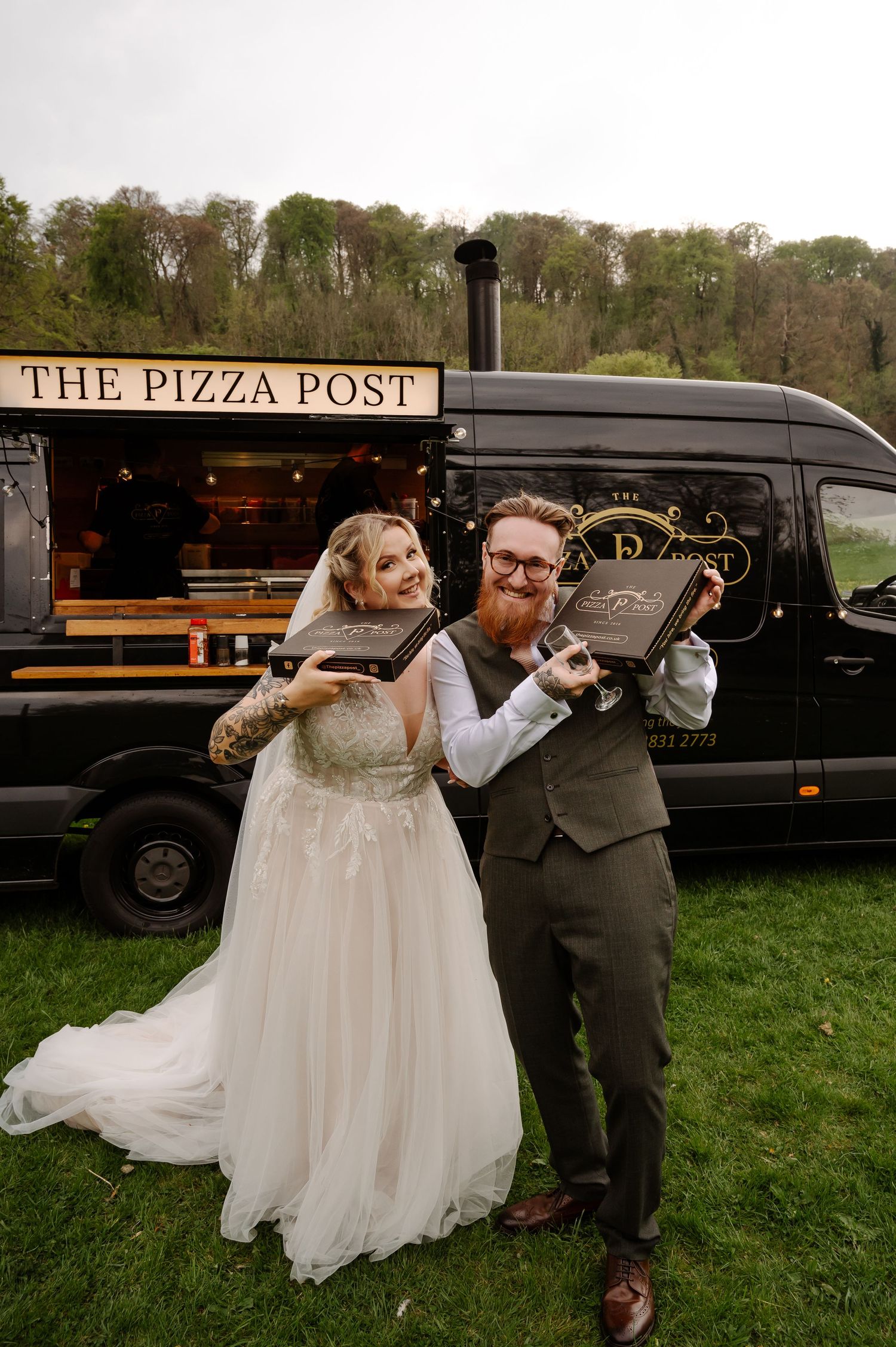A couple poses playfully with pizza boxes in front of a mobile pizza truck called The Pizza Post at an outdoor wedding reception.
