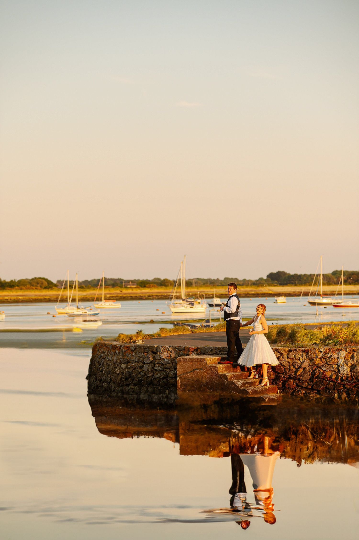 Two people in wedding attire stand on a stone jetty at sunset with sailboats moored in the calm harbor waters behind them.