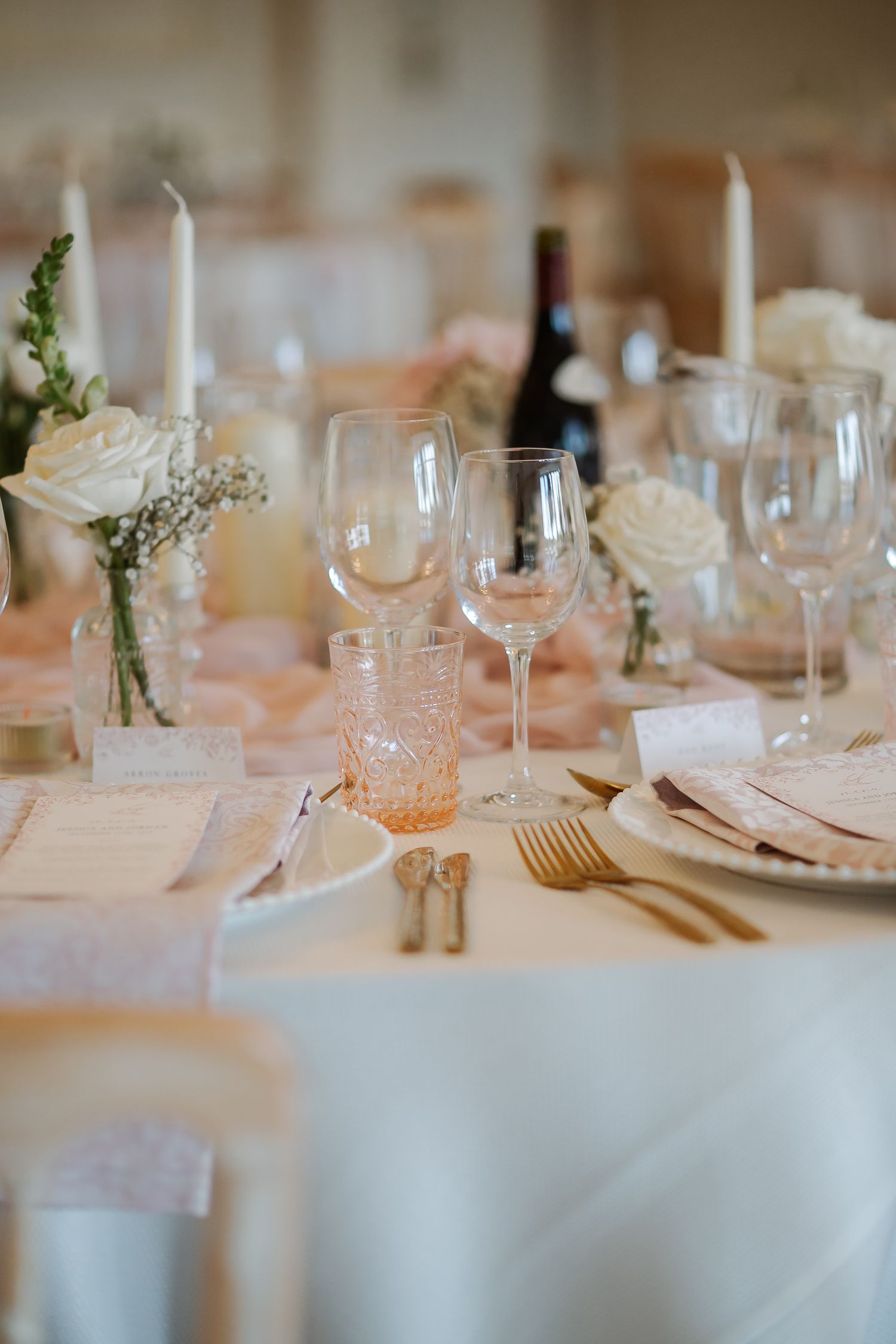 Elegant wedding table setting with white roses, gold cutlery, crystal glasses, and candles on a white linen tablecloth.