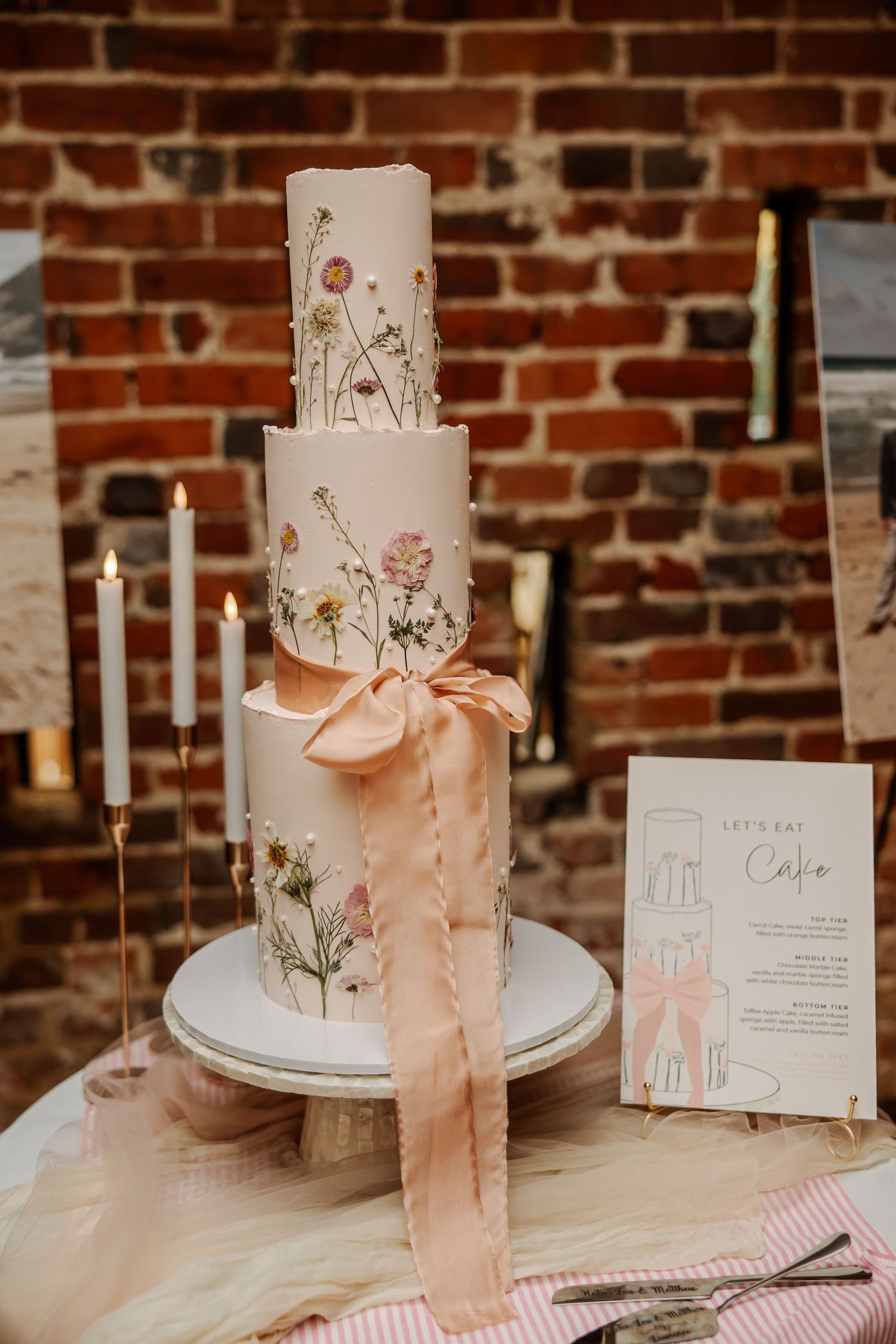 Elegant three-tier wedding cake with pressed wildflowers and pink ribbon bow, displayed near candles against a brick wall.
