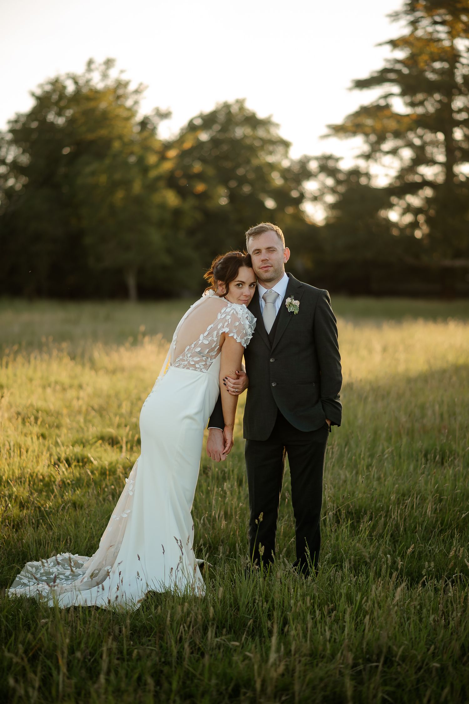 Bride in white lace gown leans on groom in dark suit during golden hour portrait in lush green field.