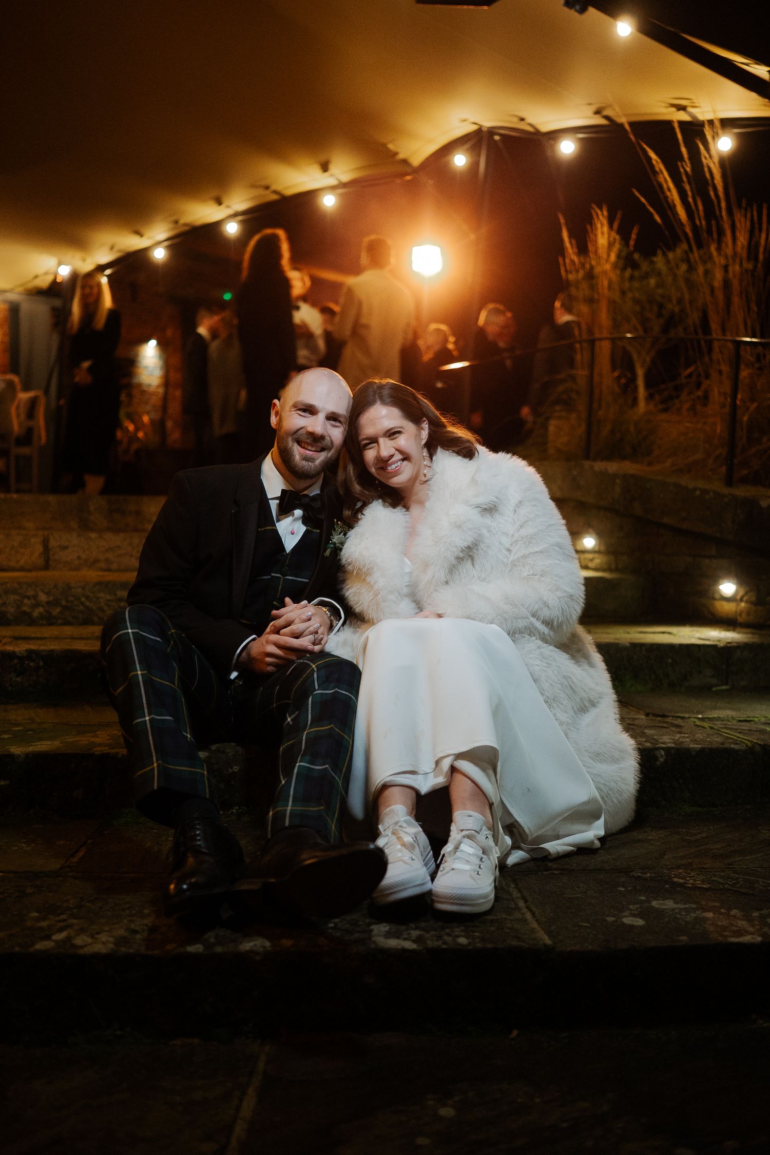 A smiling bride in a white fur coat and groom in tartan trousers sit together on steps at a night wedding reception.