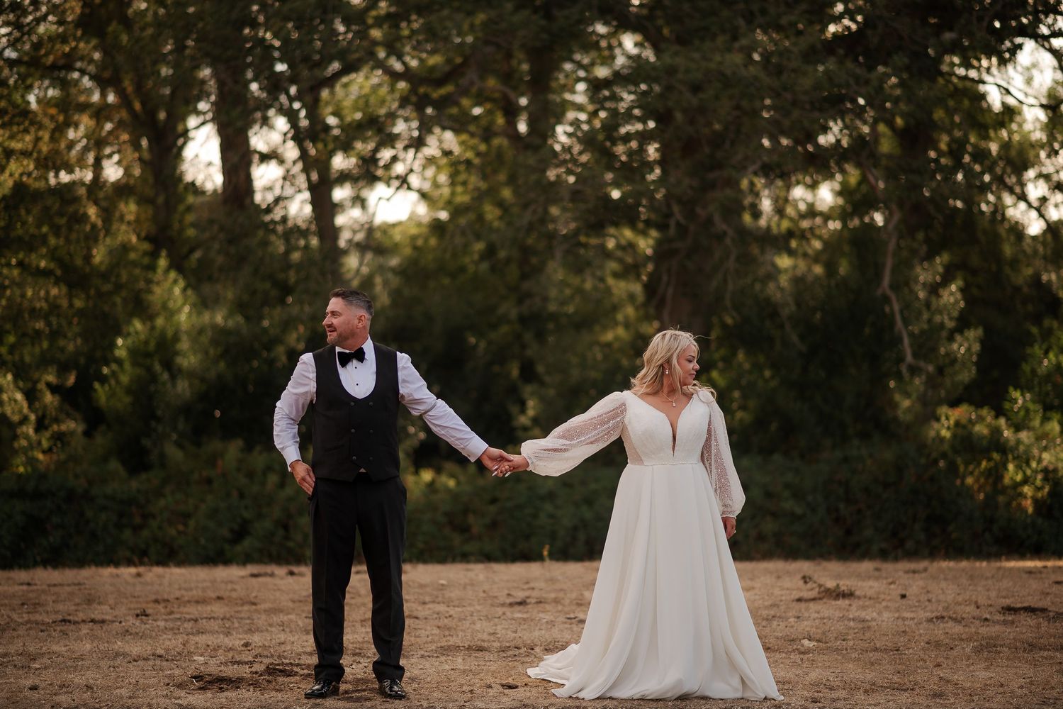 A bride and groom hold hands in a golden field surrounded by lush trees during an outdoor wedding photo session.