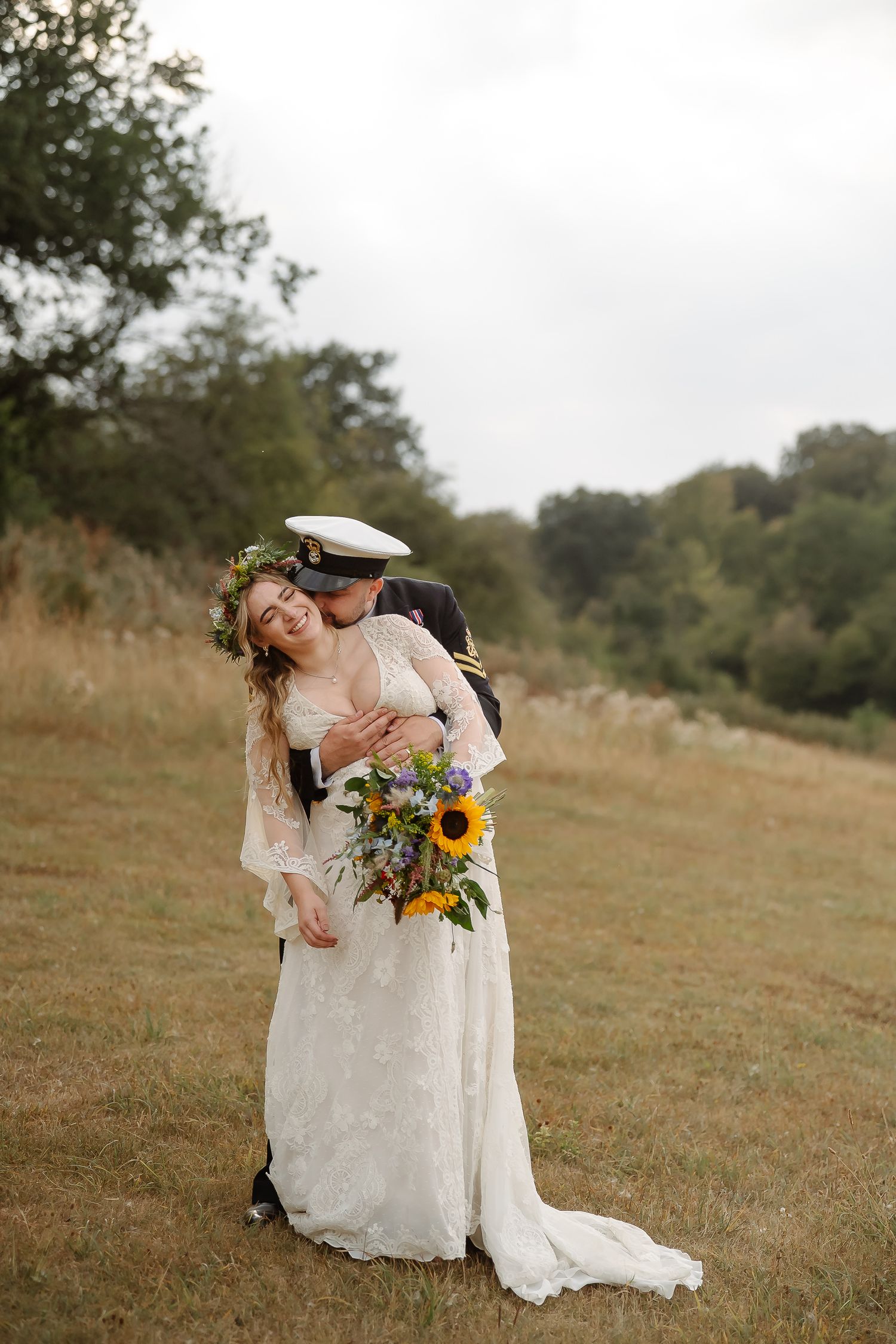 Bride in lace dress with floral crown laughs as groom in military hat embraces her outdoors in a field.