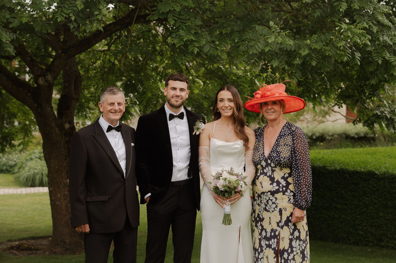 Wedding party members pose together outdoors in formal attire under green trees during a summer celebration.