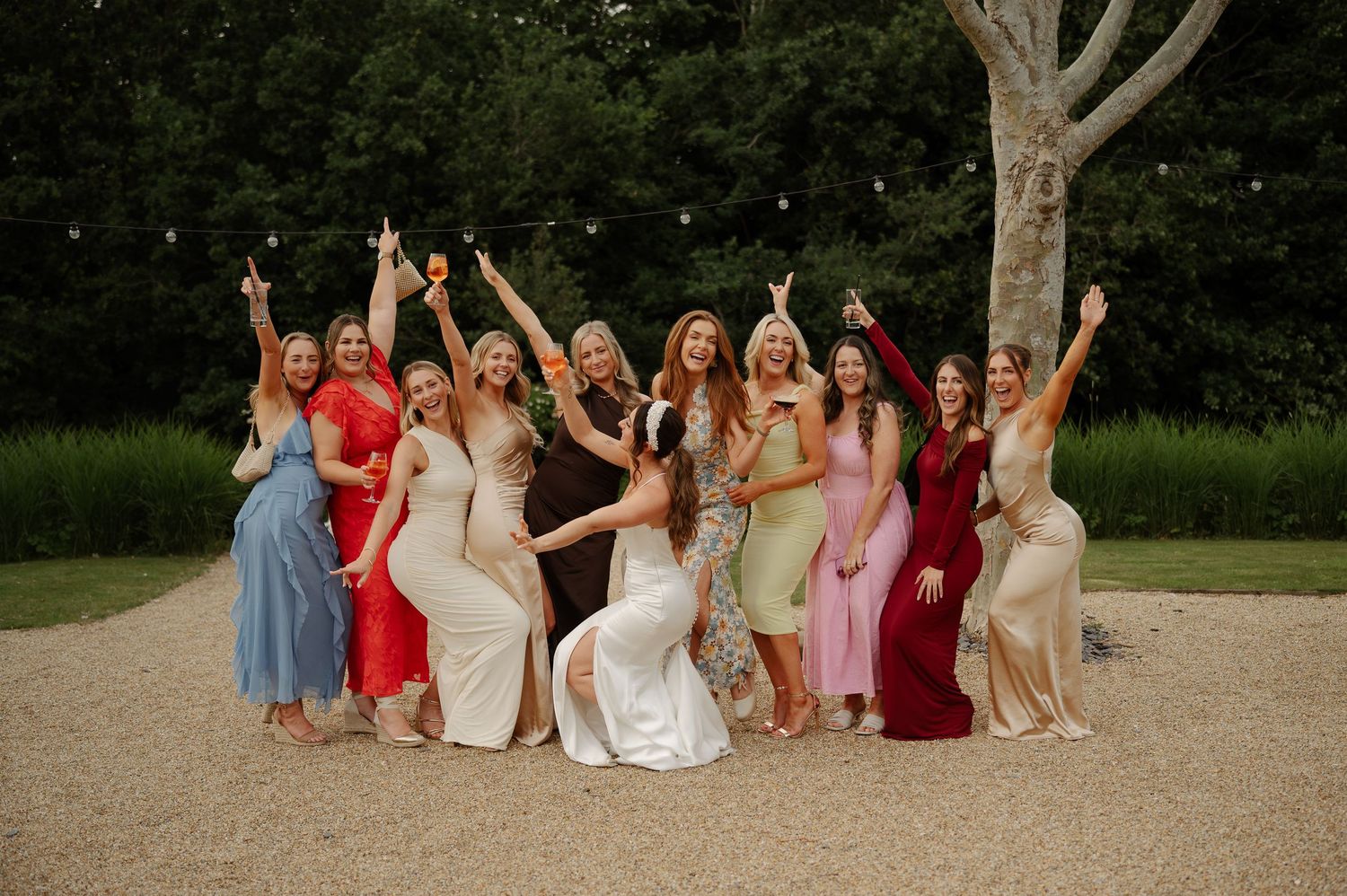 Wedding guests in colorful dresses pose playfully on the grass during outdoor celebrations.