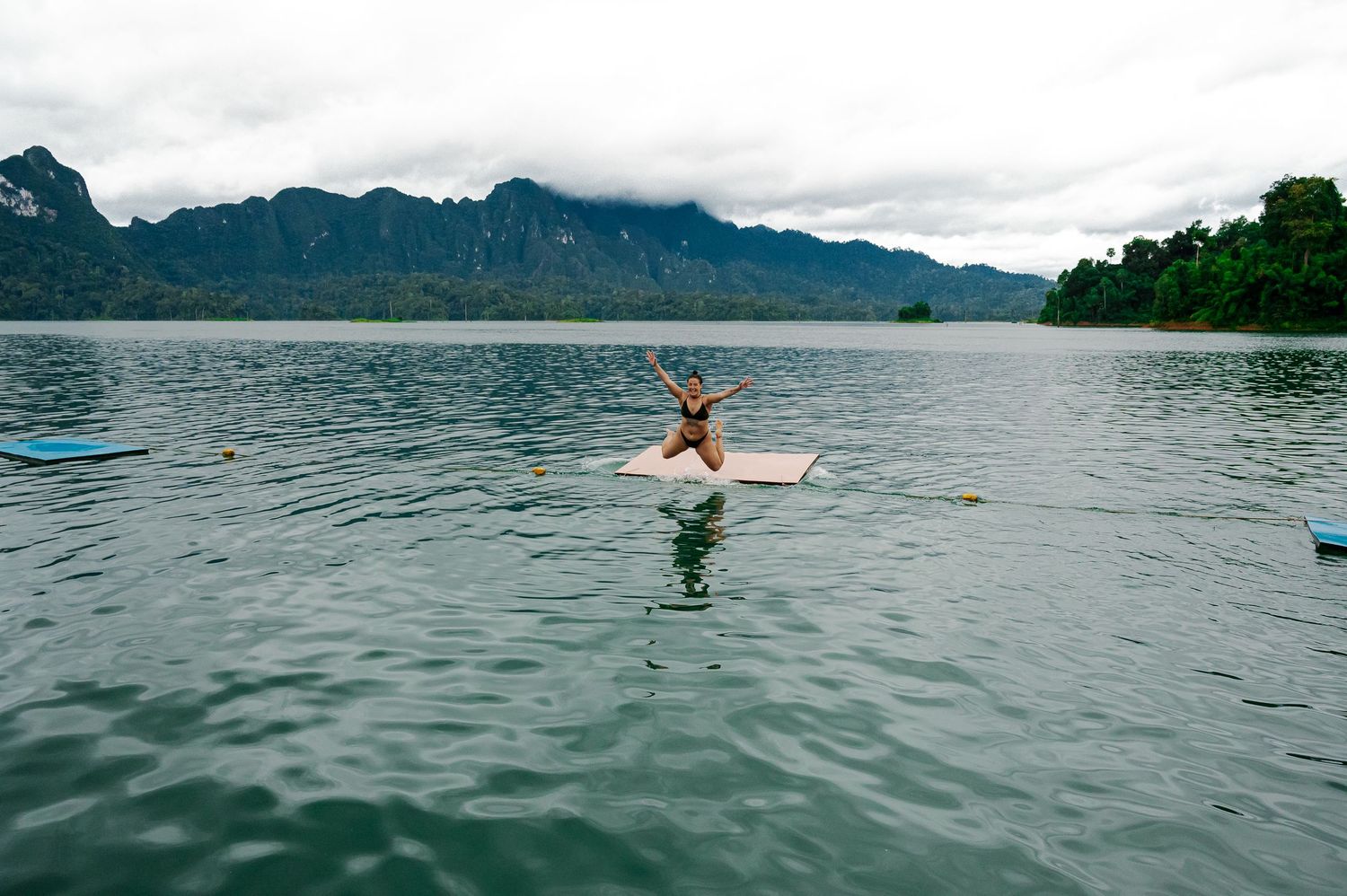 A person waterskiing on a calm lake surrounded by green mountains and forest on a cloudy day.