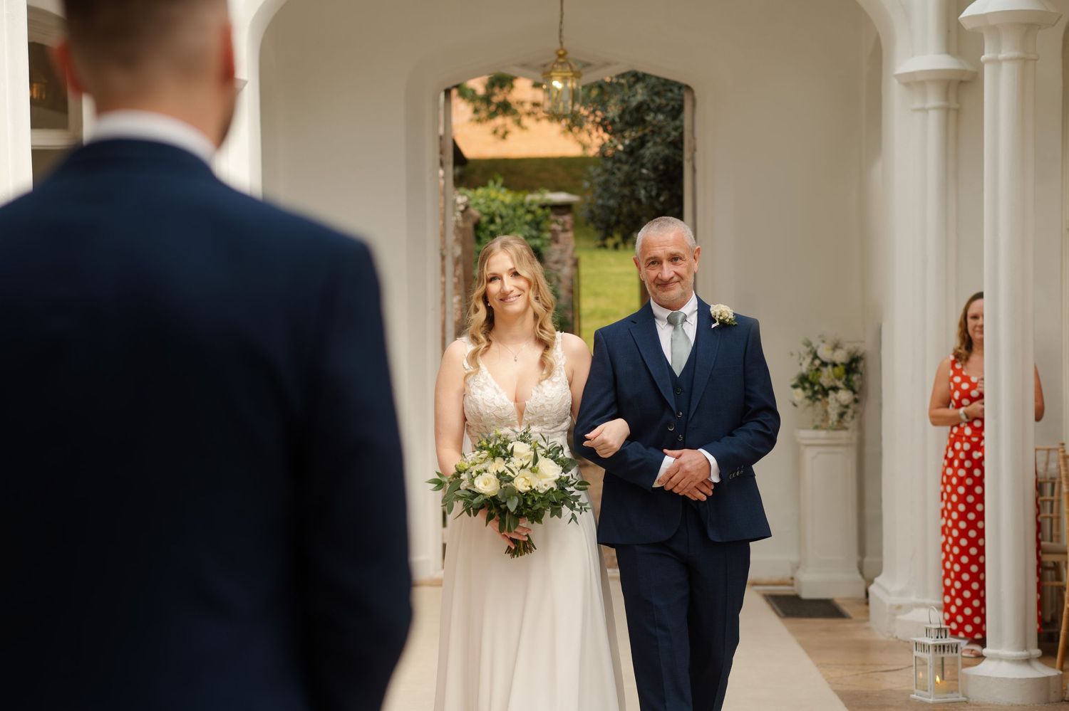 A bride in a white gown holds a floral bouquet while walking down a columned hallway with someone in a navy suit.