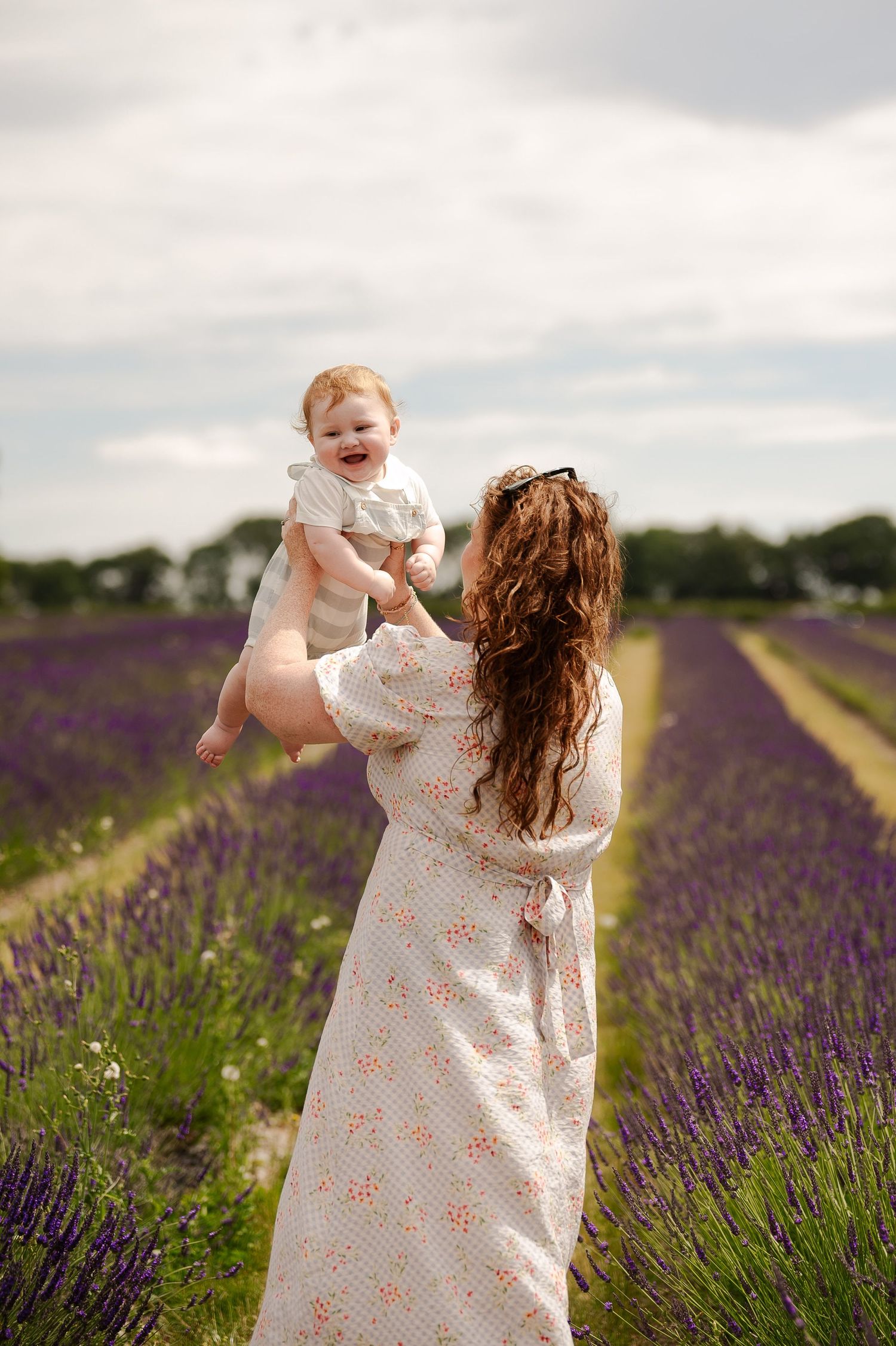 A person in a floral dress holds a baby while standing in a beautiful purple lavender field on a cloudy summer day.