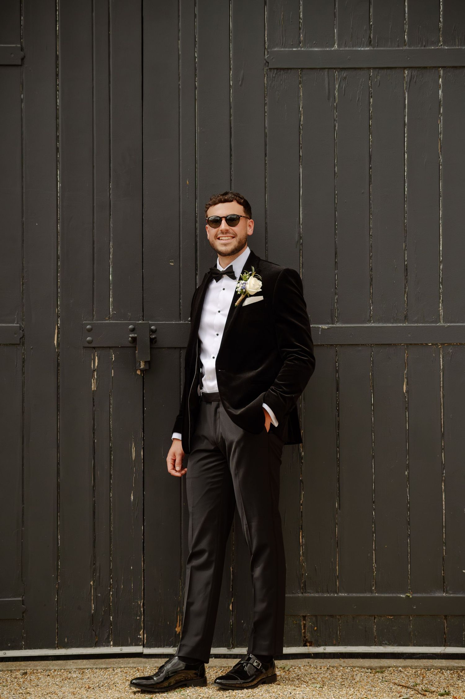 Formal wedding party members in black tuxedos pose against a dark wooden door backdrop.