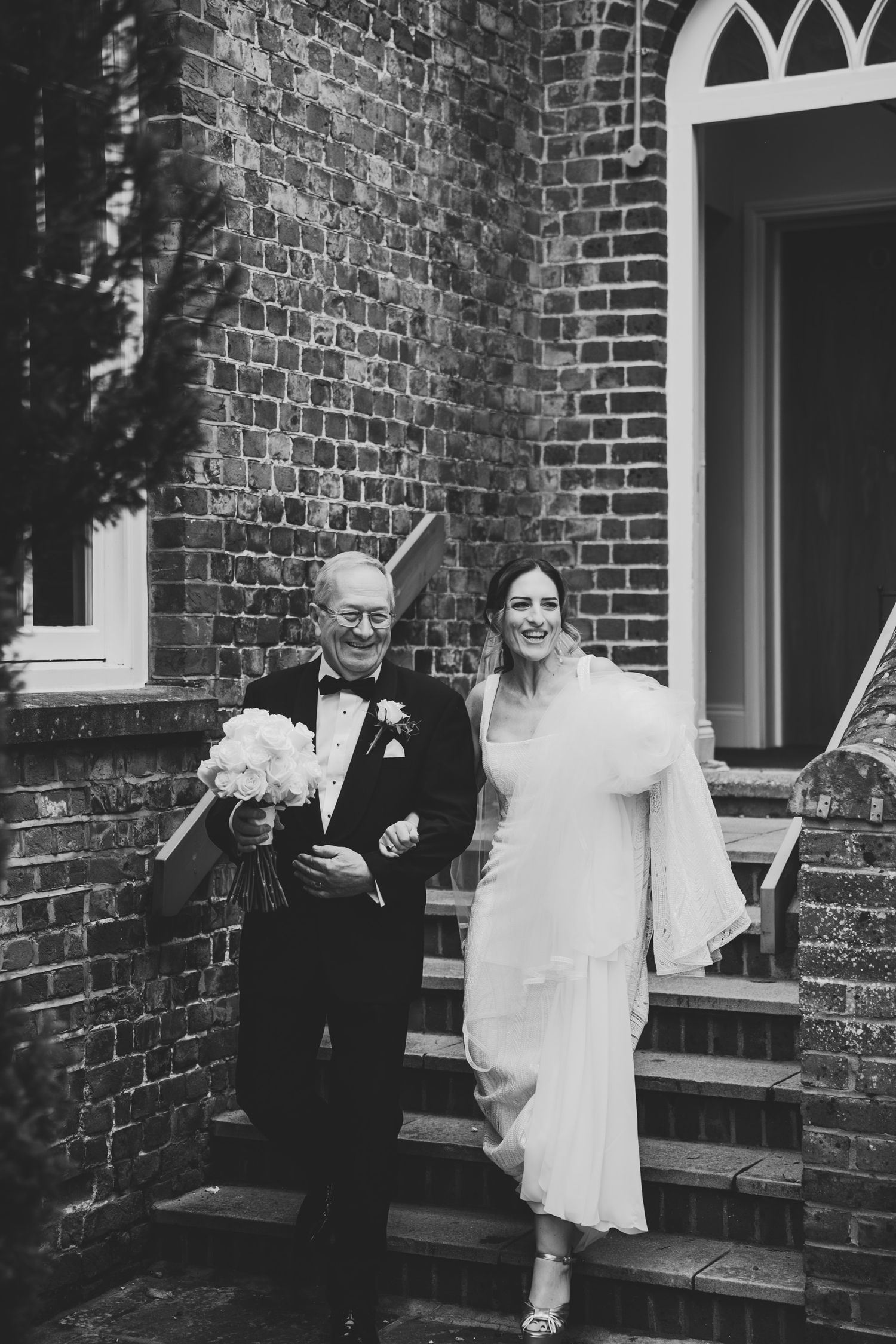 Bride in white gown walks with elderly man in tuxedo outside brick church in elegant black and white wedding photo.