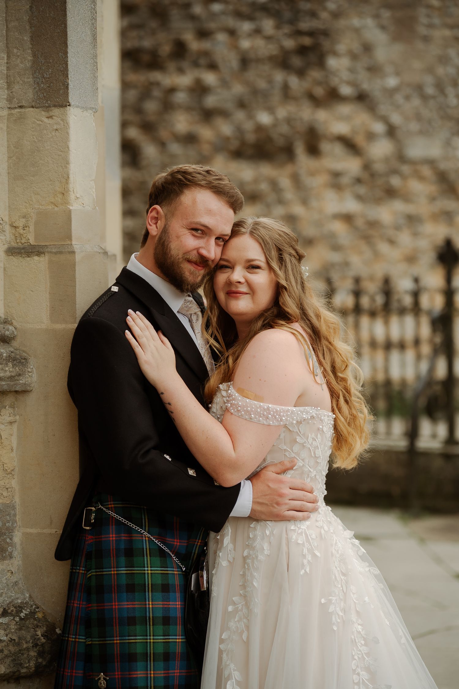 A couple in wedding attire embraces outdoors, groom wearing a tartan kilt and bride in an off-shoulder lace gown.