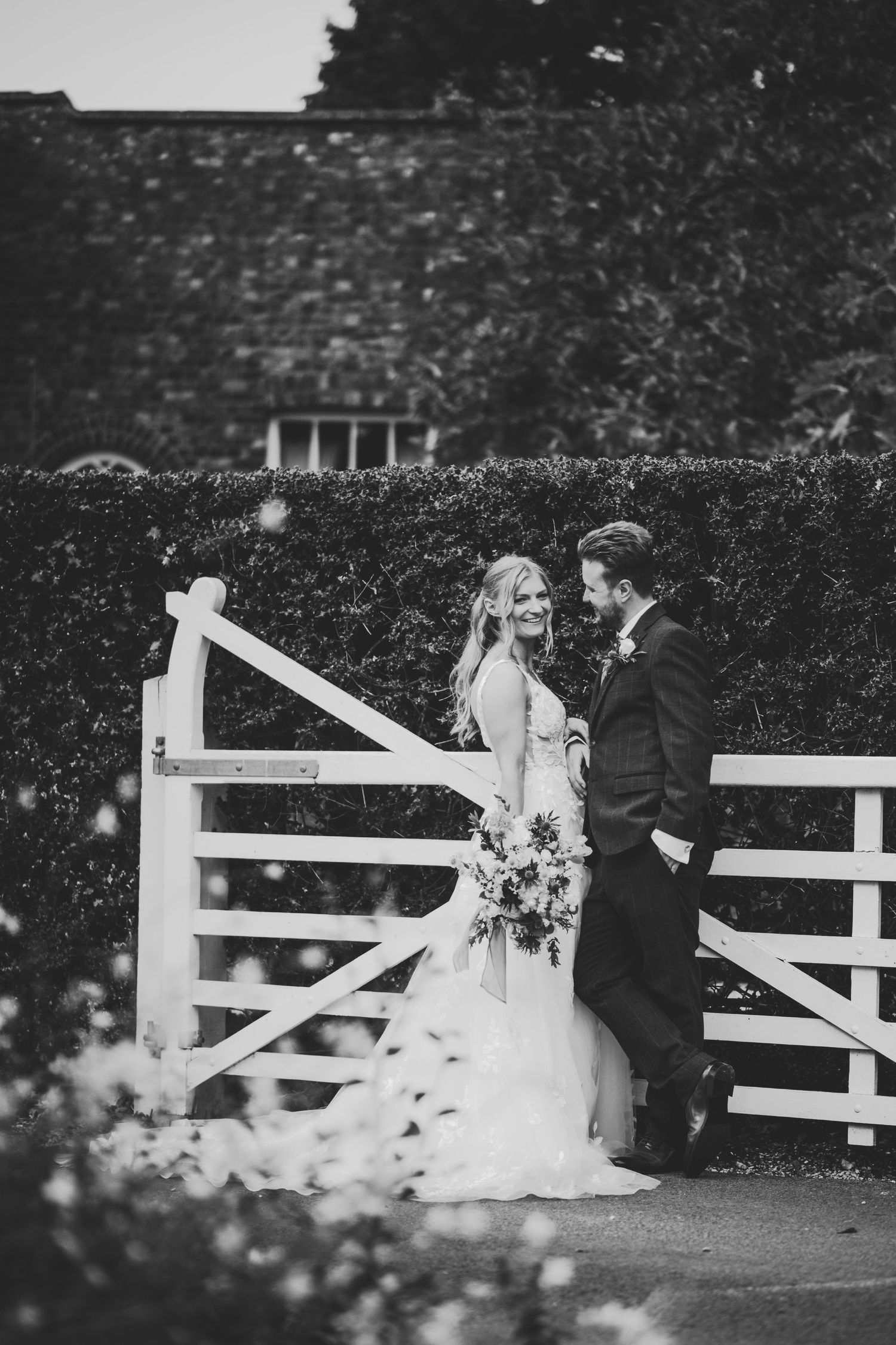 Black and white photo of a bride and groom sharing a romantic moment by a white wooden gate at a countryside wedding.