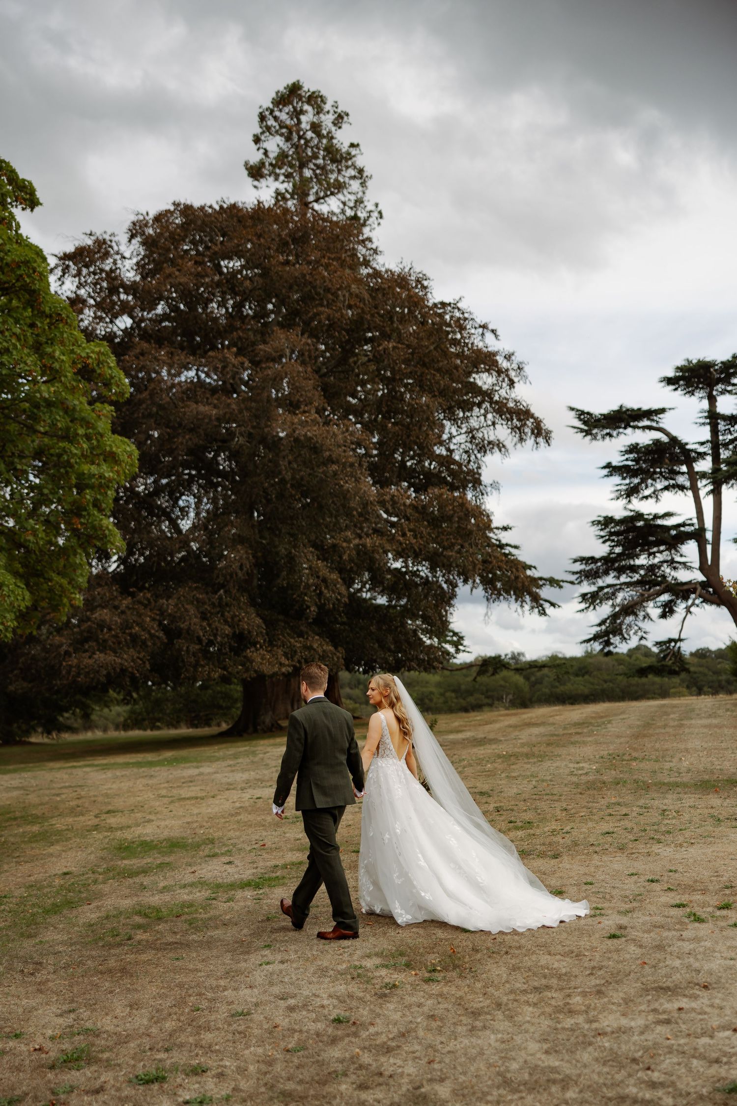Bride and groom walk hand in hand across a grassy estate, her flowing white gown and veil trailing behind them.