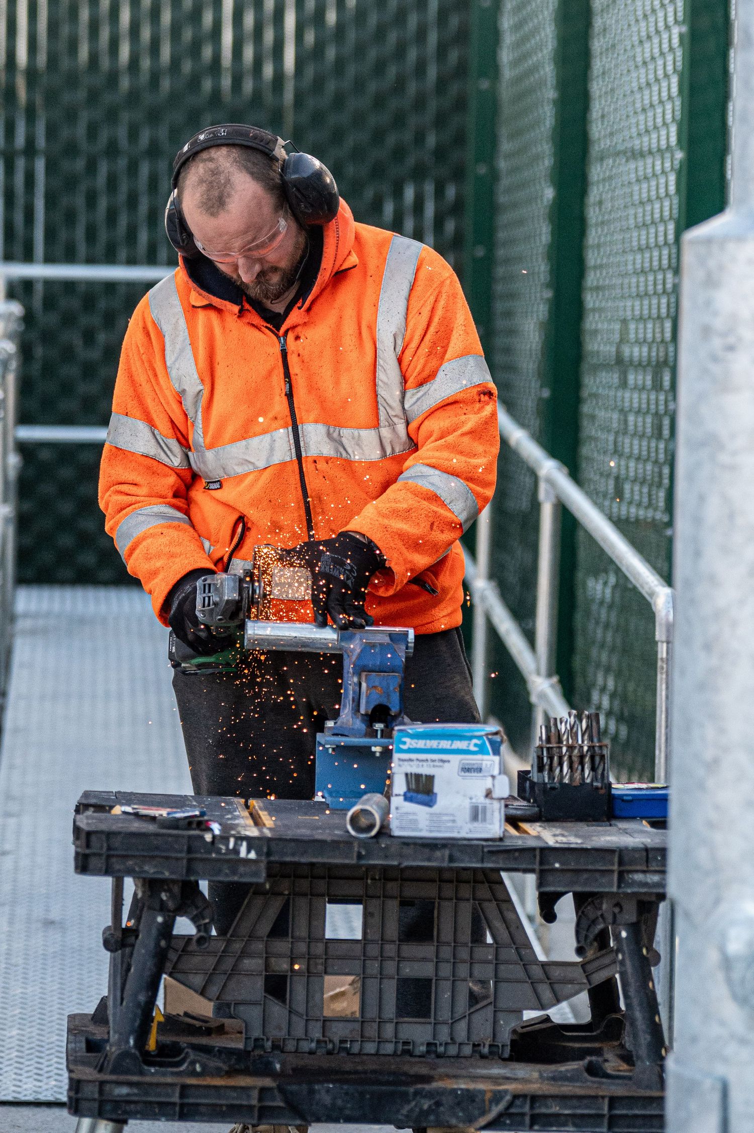 Construction worker in high visibility orange jacket operates power tools on a workbench with sparks flying.