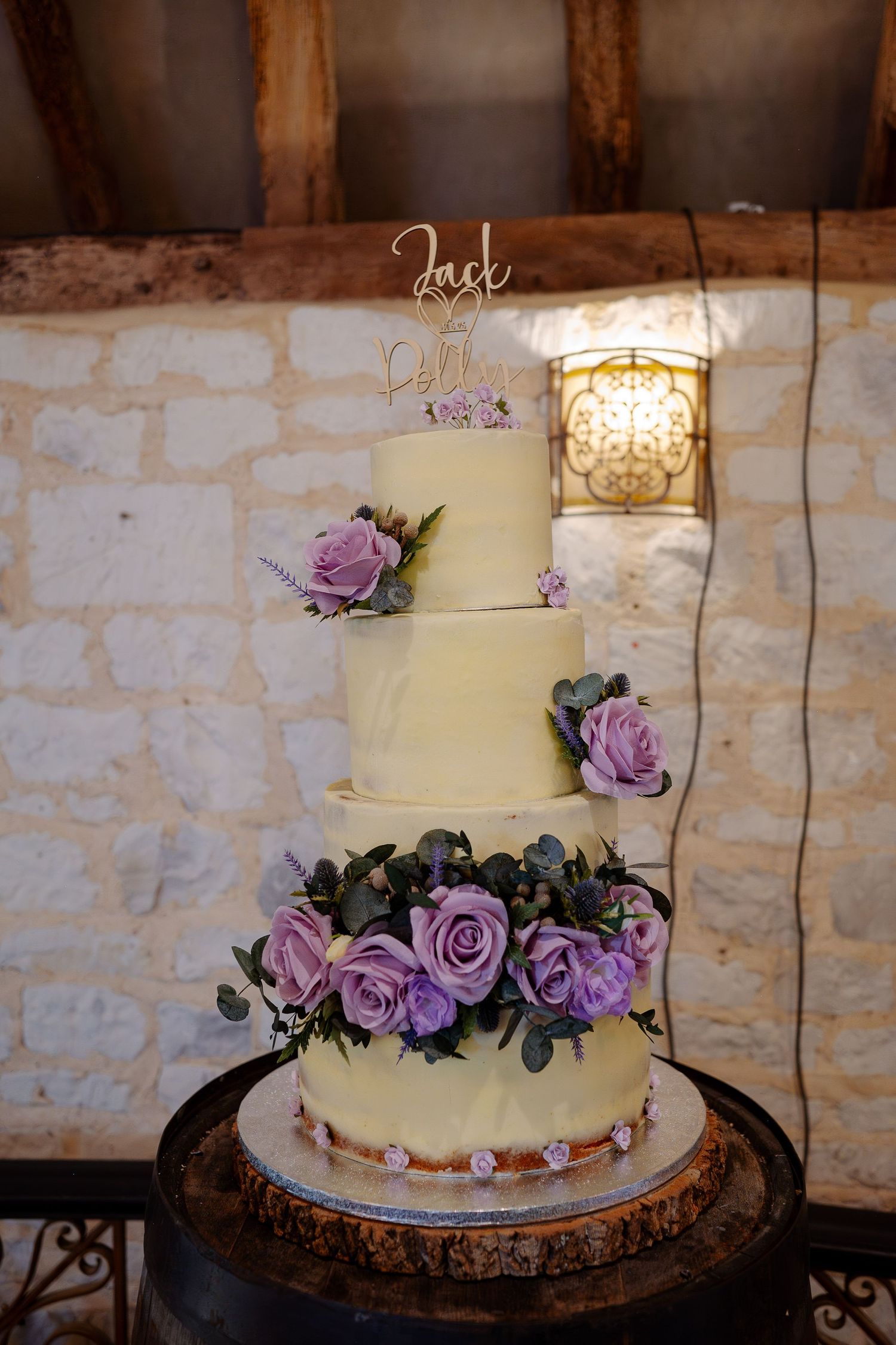 Three-tiered white wedding cake adorned with purple roses and delicate floral decorations on a rustic stand.