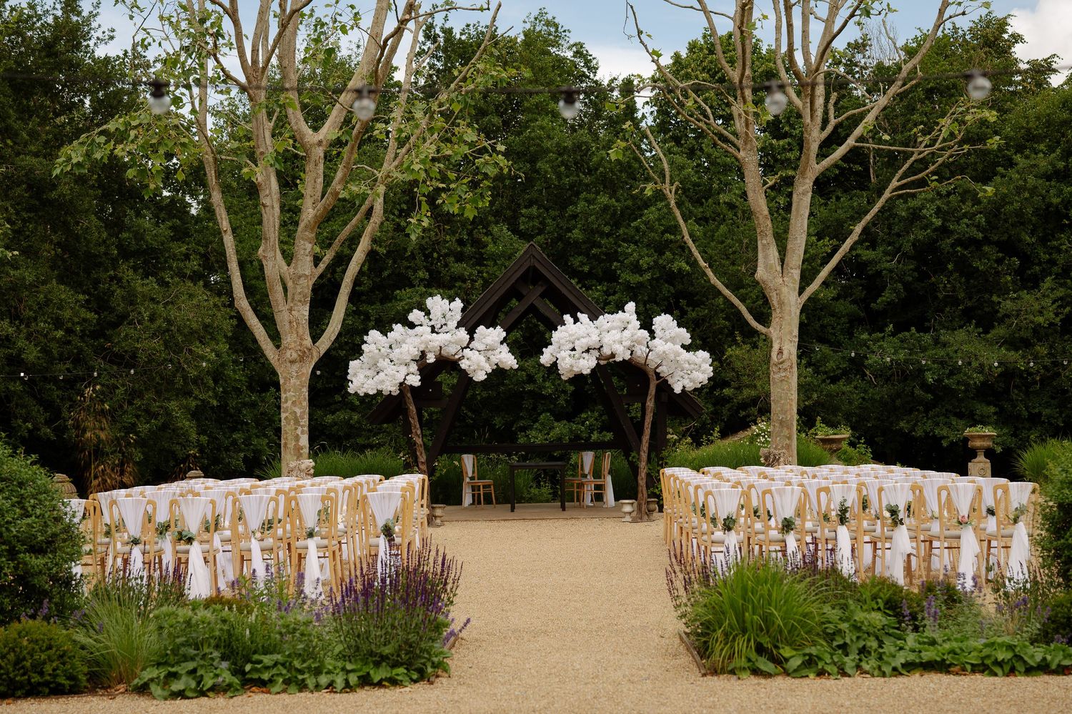 Panoramic view of outdoor wedding ceremony setup with guests seated in rows among trees and gardens.