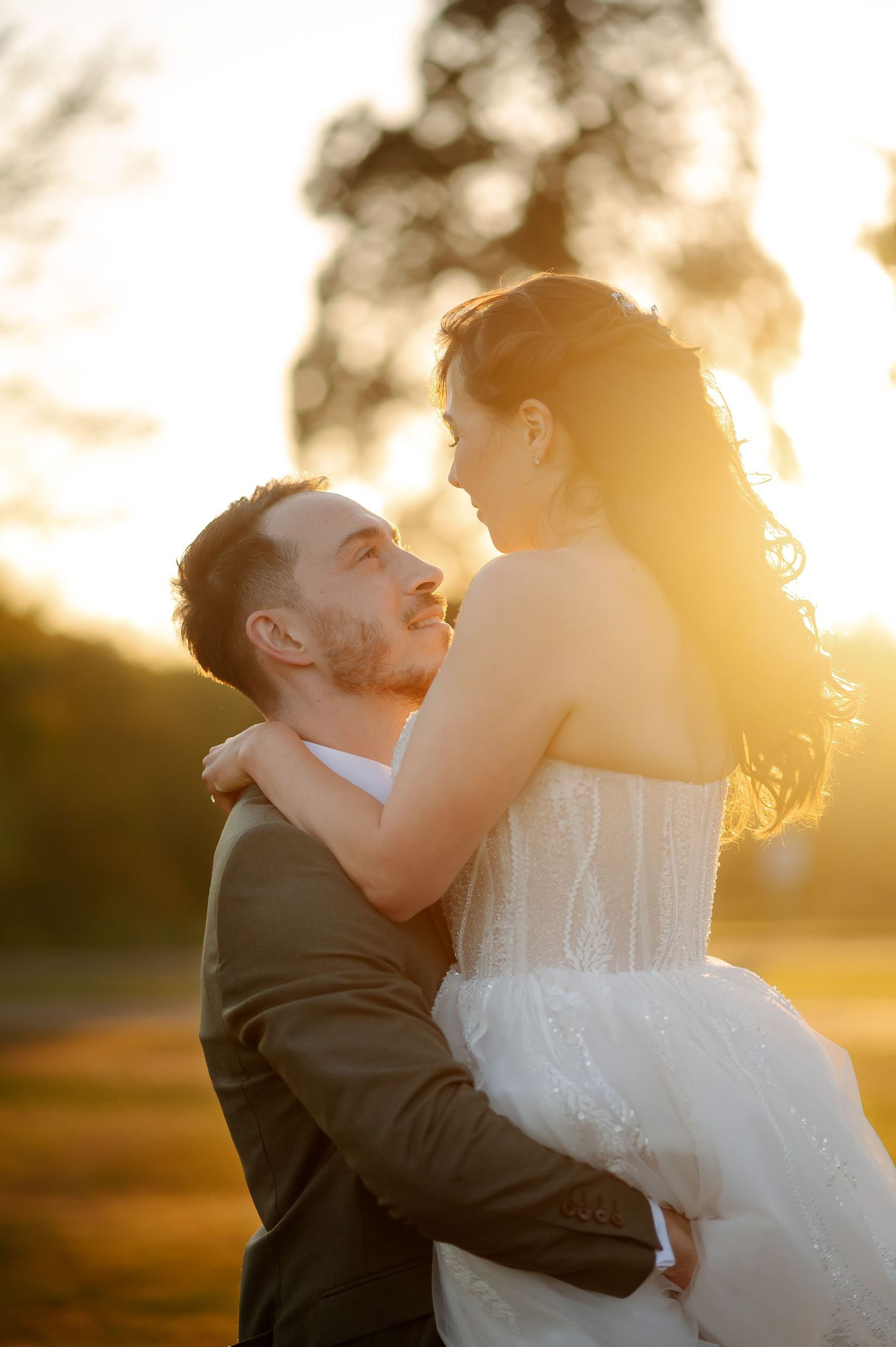 A couple shares a romantic golden hour embrace outdoors, the groom lifting his bride in a white gown at sunset.